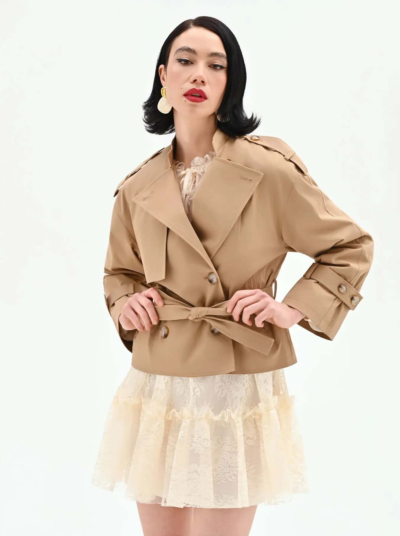 A woman with black hair poses confidently in a beige trench coat over a cream lace dress. She wears large hoop earrings and bold red lipstick.