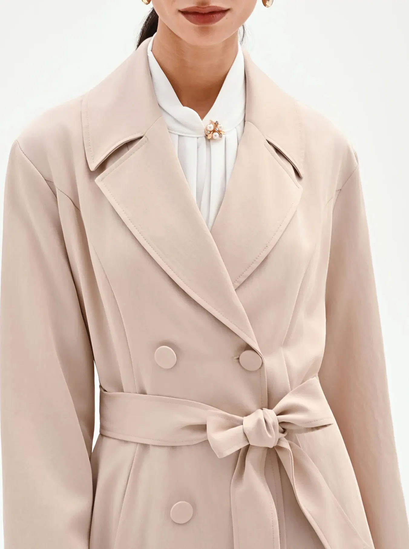 A woman models a stylish beige trench coat over wide-leg plaid pants. Her hair is neatly tied back, and she exudes a sophisticated, confident demeanor.