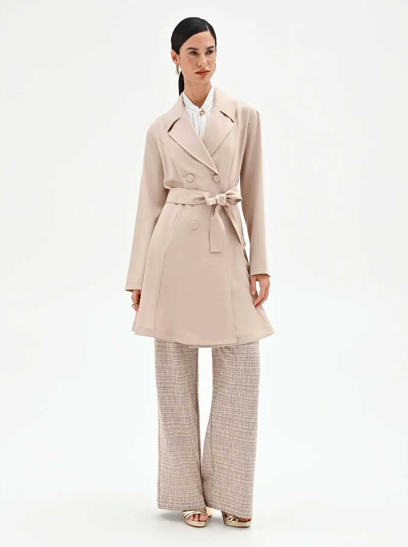 A woman models a stylish beige trench coat over wide-leg plaid pants. Her hair is neatly tied back, and she exudes a sophisticated, confident demeanor.