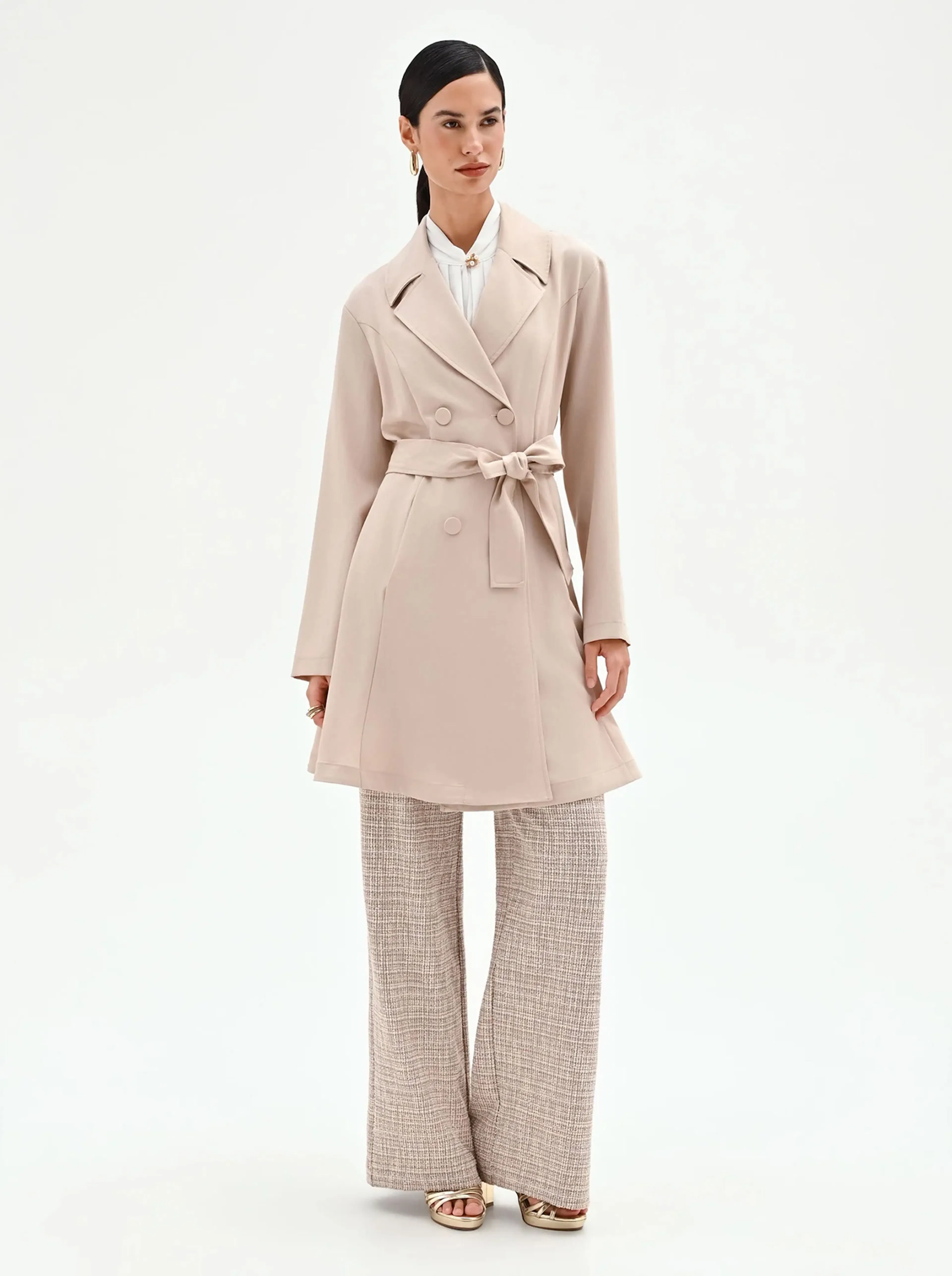 A woman models a stylish beige trench coat over wide-leg plaid pants. Her hair is neatly tied back, and she exudes a sophisticated, confident demeanor.