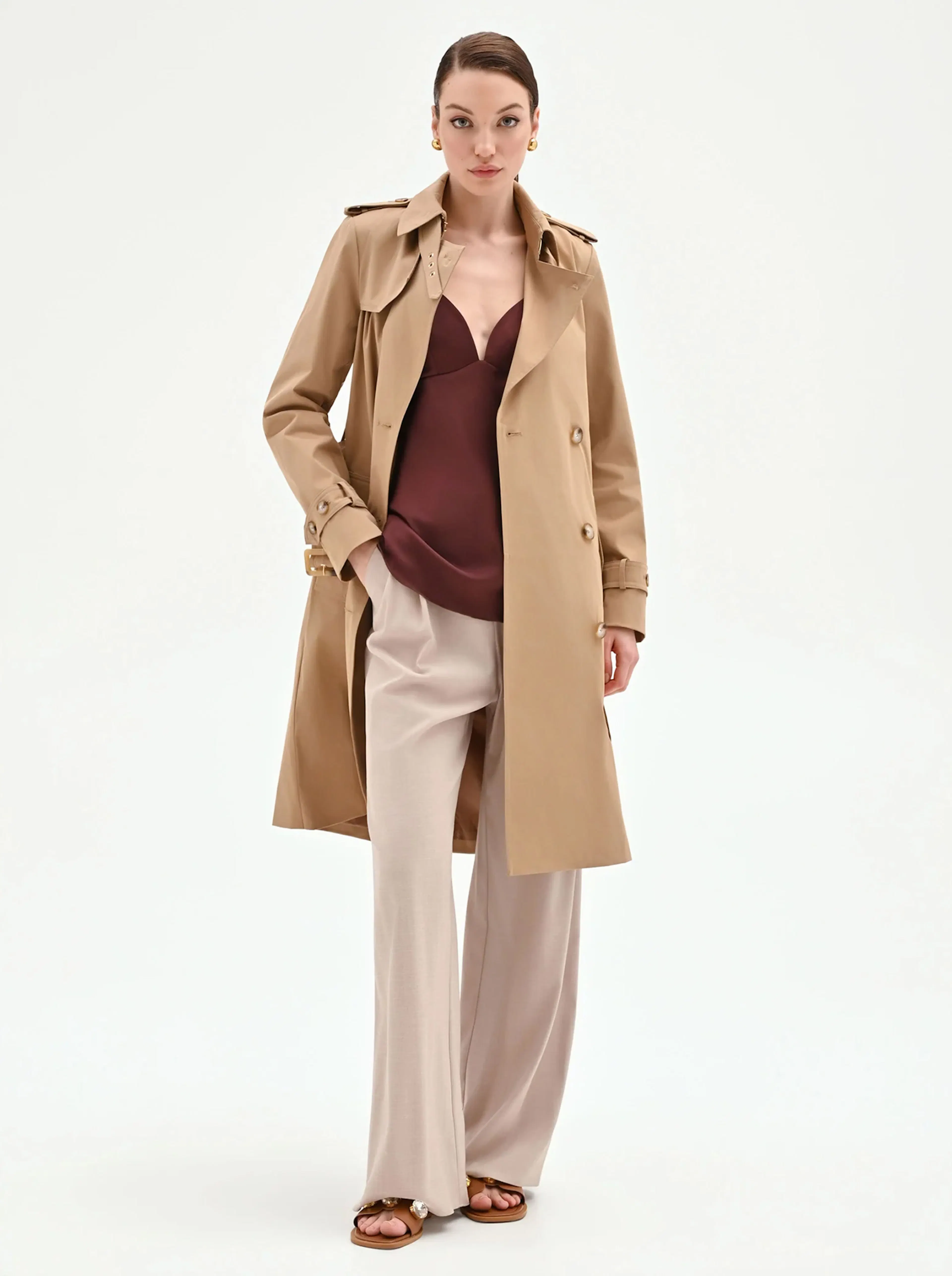 A model wearing a beige trench coat over a burgundy top and cream wide-leg pants. The look is elegant and sophisticated, complemented by gold earrings.