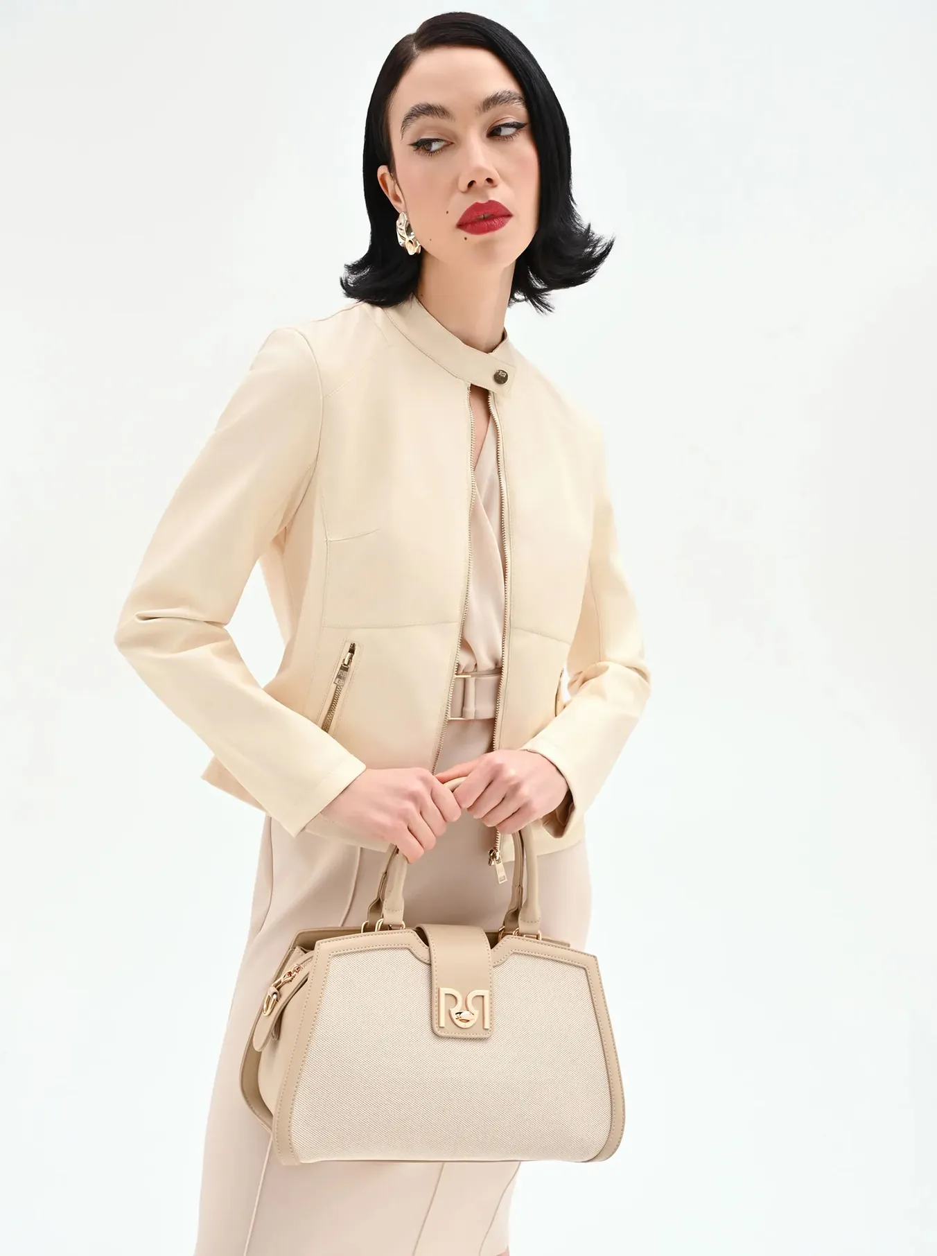Woman in elegant beige attire with a matching handbag stands against a white background. Her expression is poised with bold red lipstick.