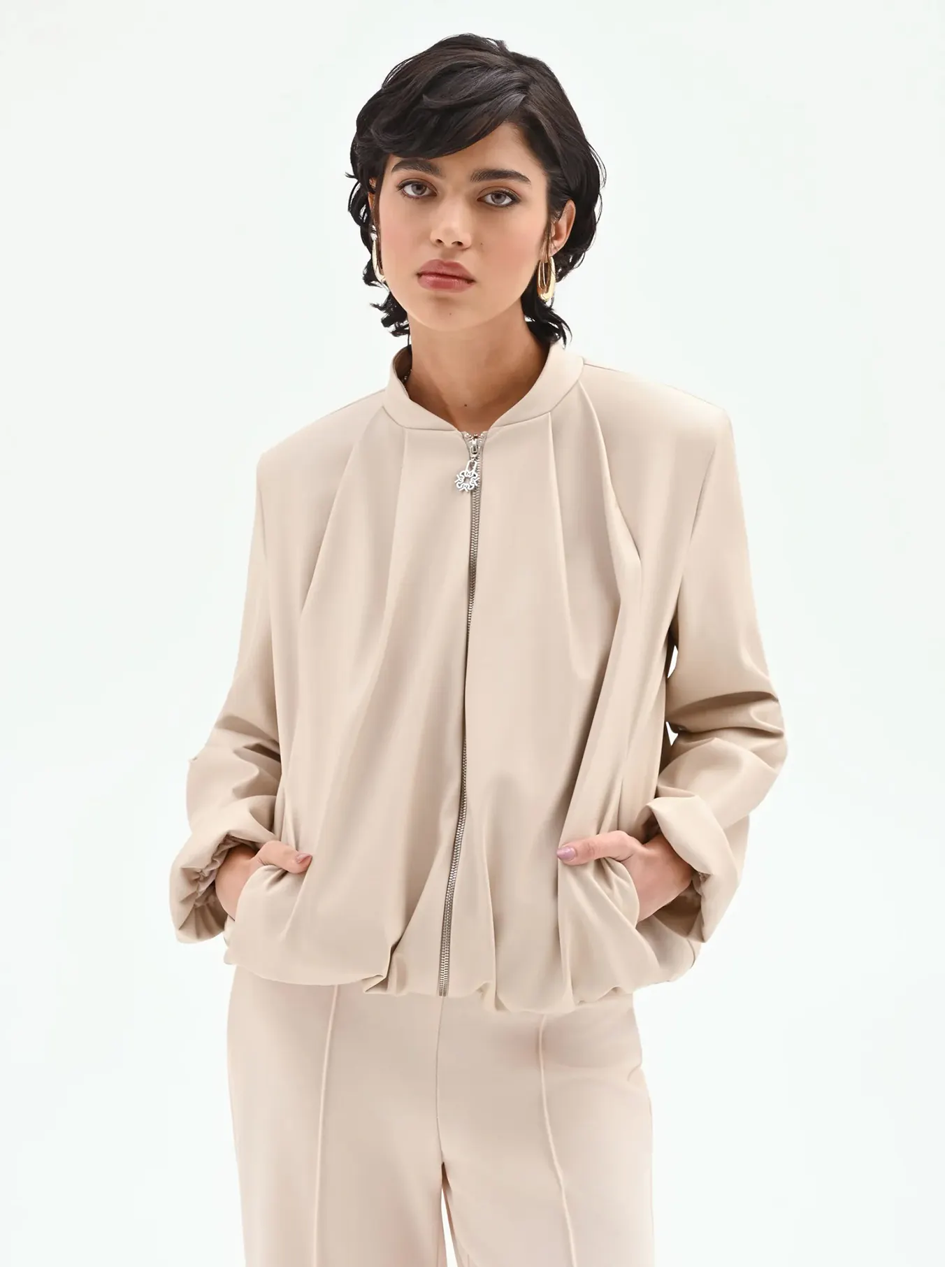 A person with short dark hair wears a beige zip-up jacket and matching trousers. They have a calm expression, hands in the jacket pockets, against a white background.