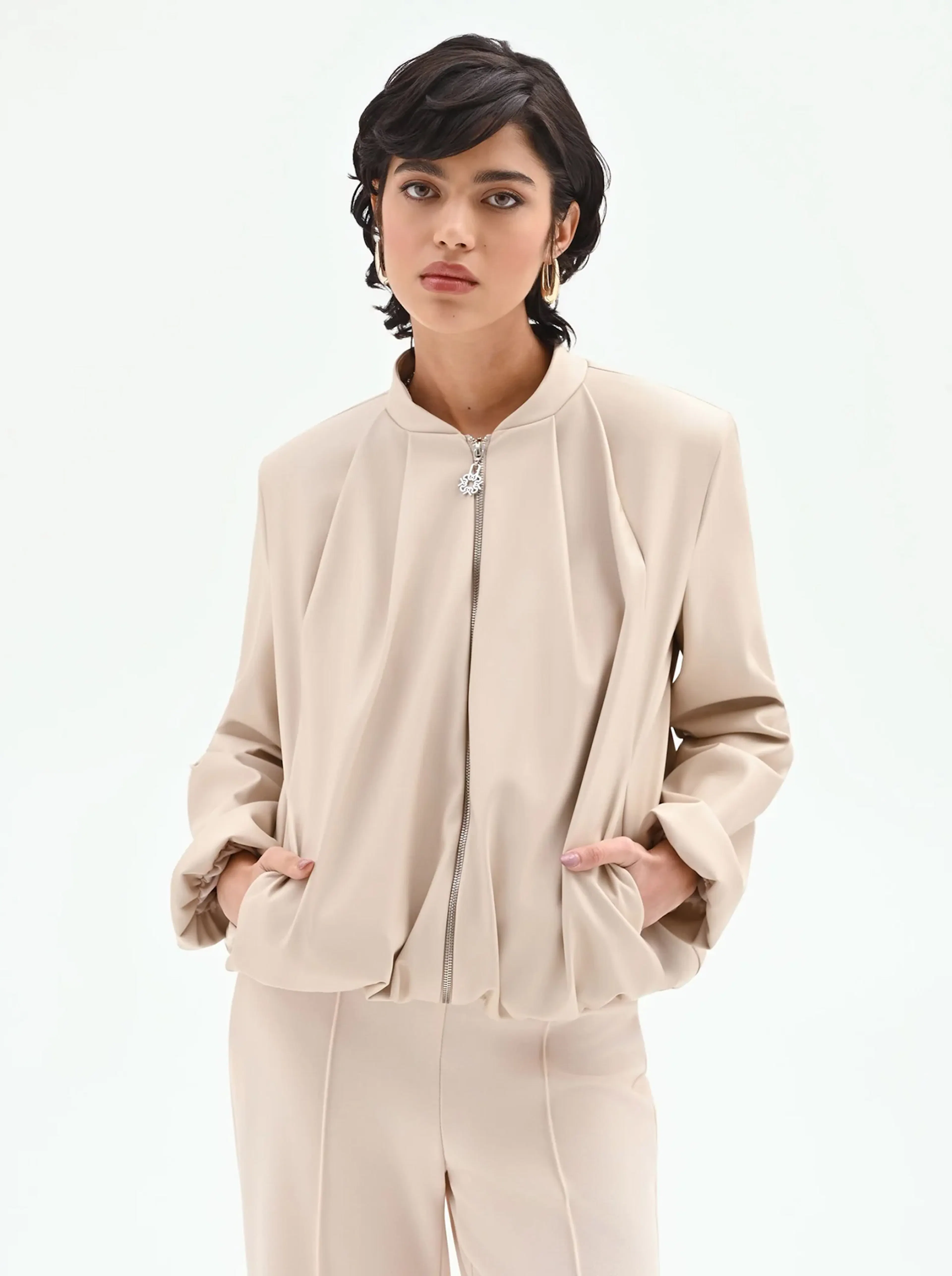 A person with short dark hair wears a beige zip-up jacket and matching trousers. They have a calm expression, hands in the jacket pockets, against a white background.