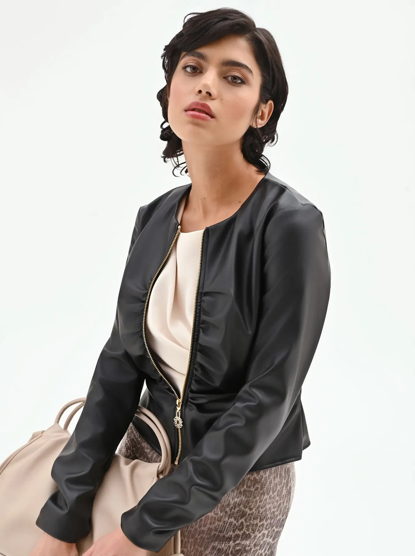 Woman in a black zip-up jacket, cream top, and patterned skirt holding a beige handbag against a plain white background.