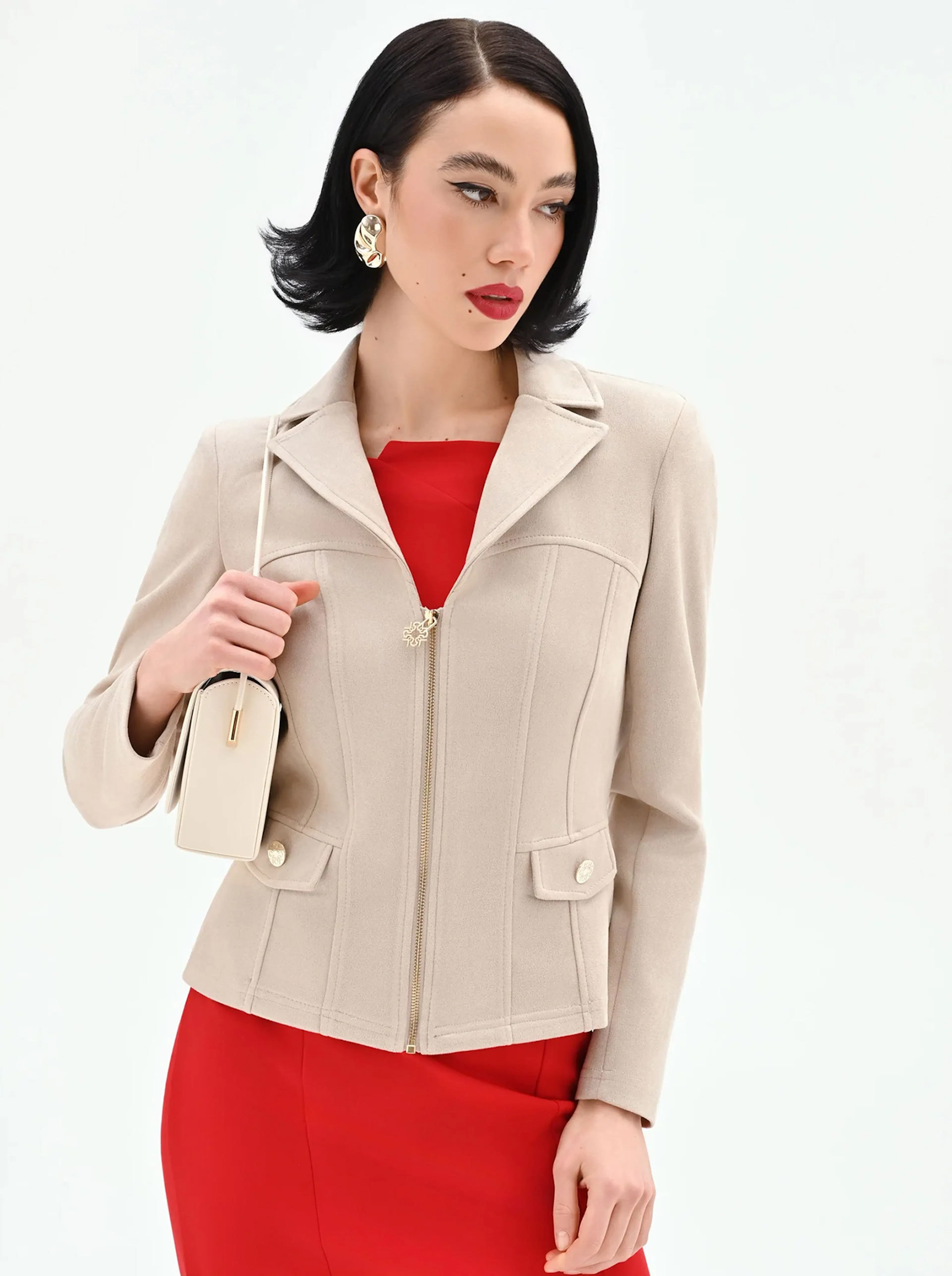 Elegant woman in a beige blazer and red dress. She holds a small beige purse and wears statement earrings. Her expression is poised and confident.