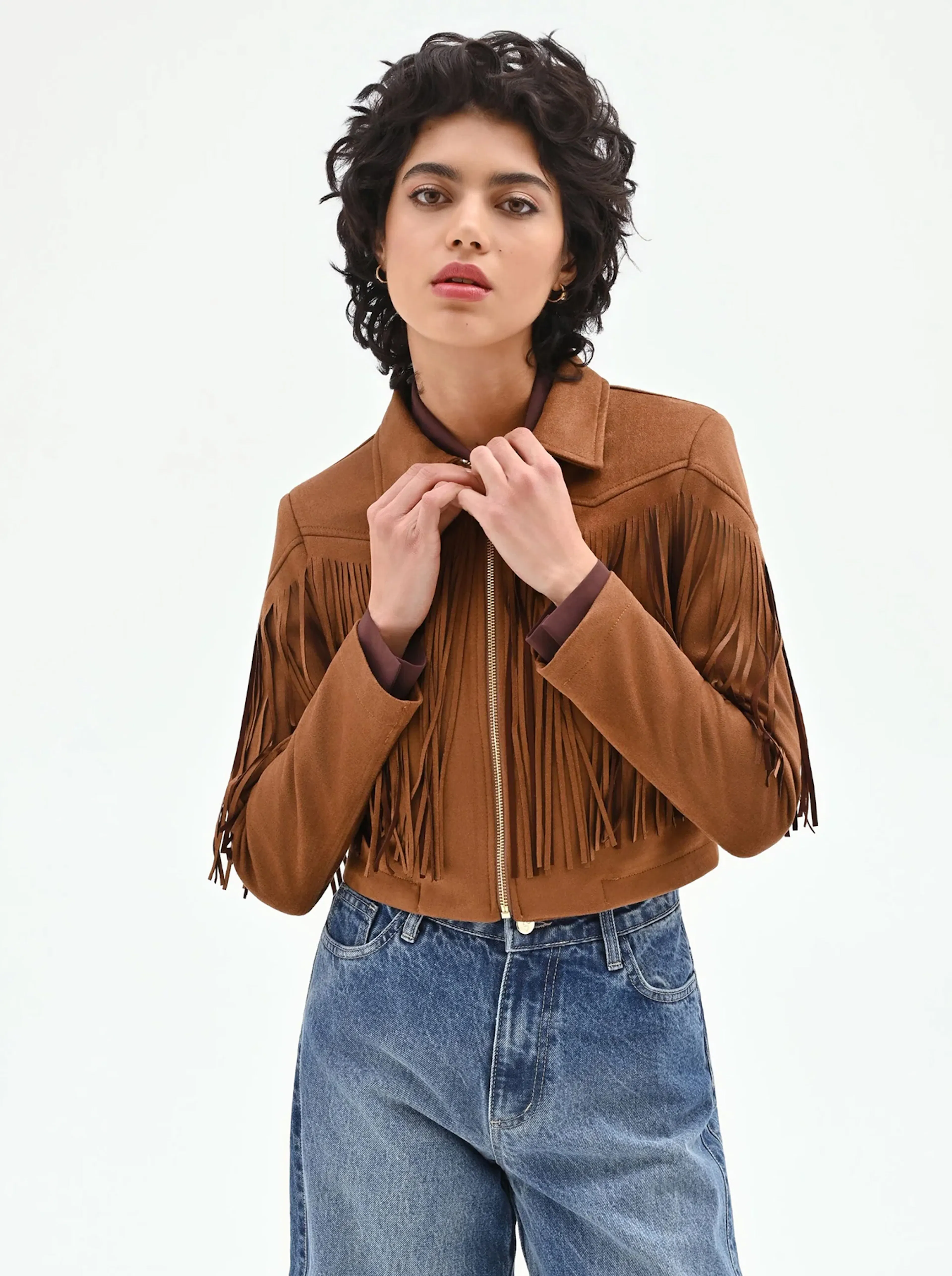 Person wearing a brown suede fringe jacket over a dark shirt and high-waisted blue jeans, adjusting the jacket collar.
