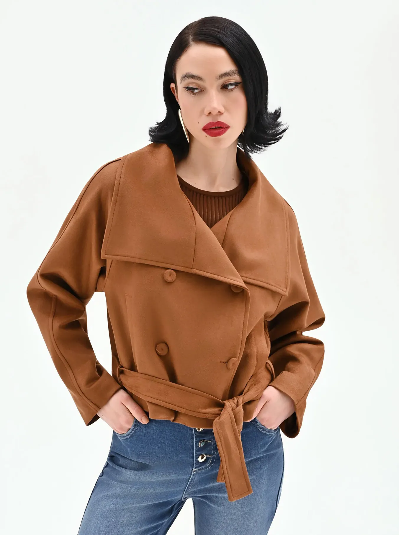 Person wearing a brown belted double-breasted jacket and blue jeans, standing with hands in pockets against a plain white background.