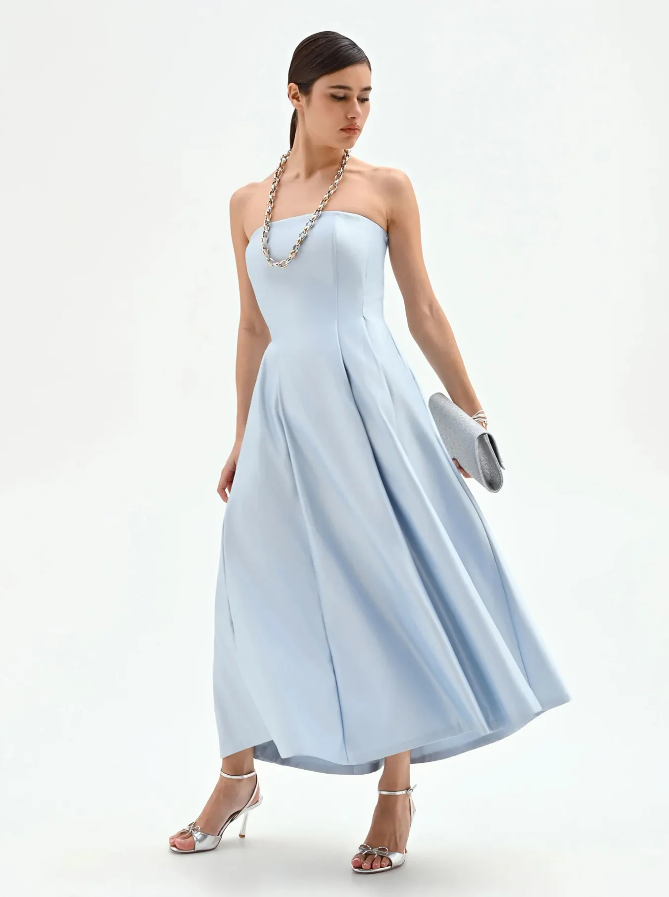 Woman wearing a strapless light blue midi dress with a silver chain necklace, silver clutch, and silver high-heeled sandals, posing against white background.