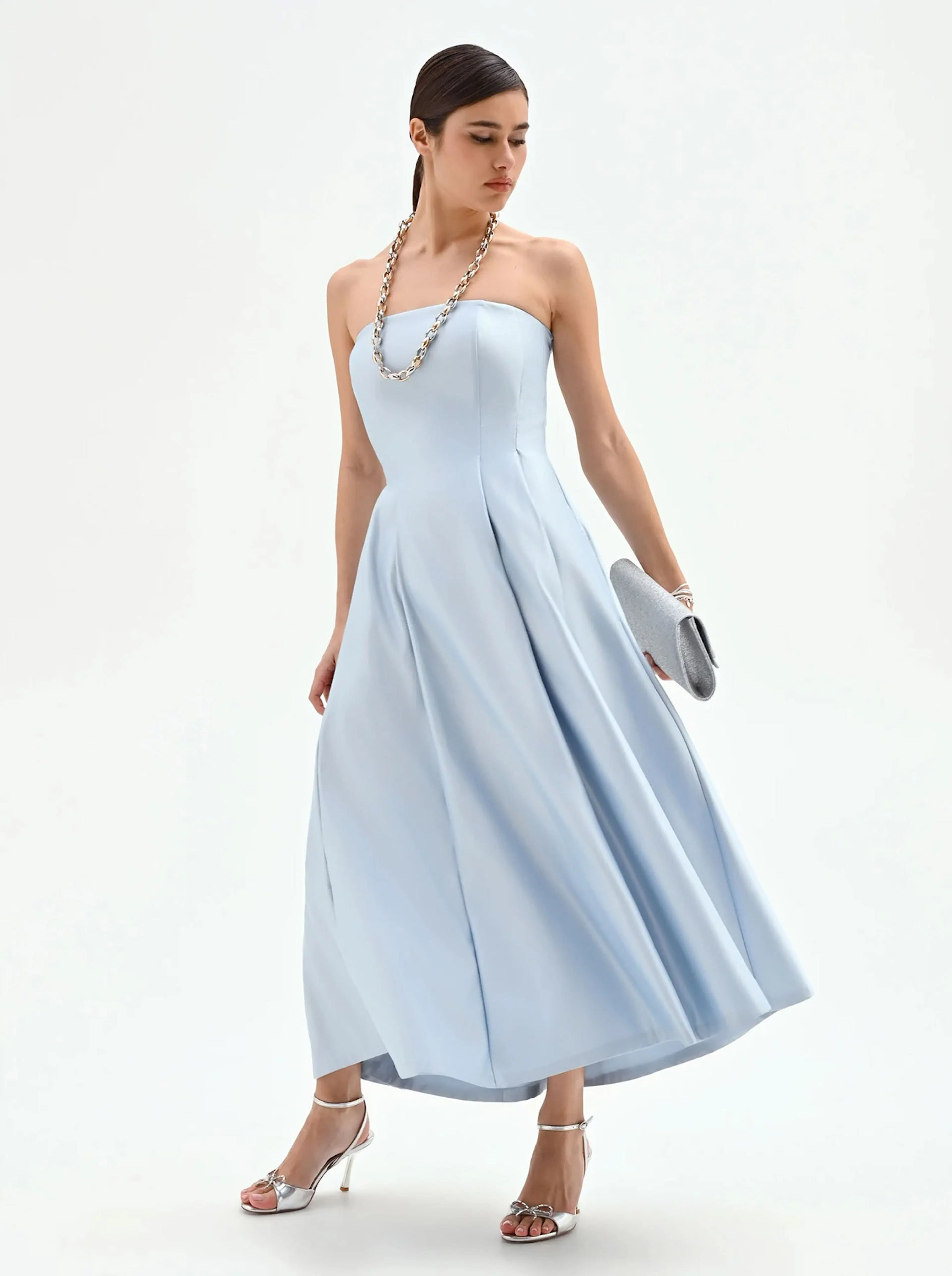 Woman wearing a strapless light blue midi dress with a silver chain necklace, silver clutch, and silver high-heeled sandals, posing against white background.
