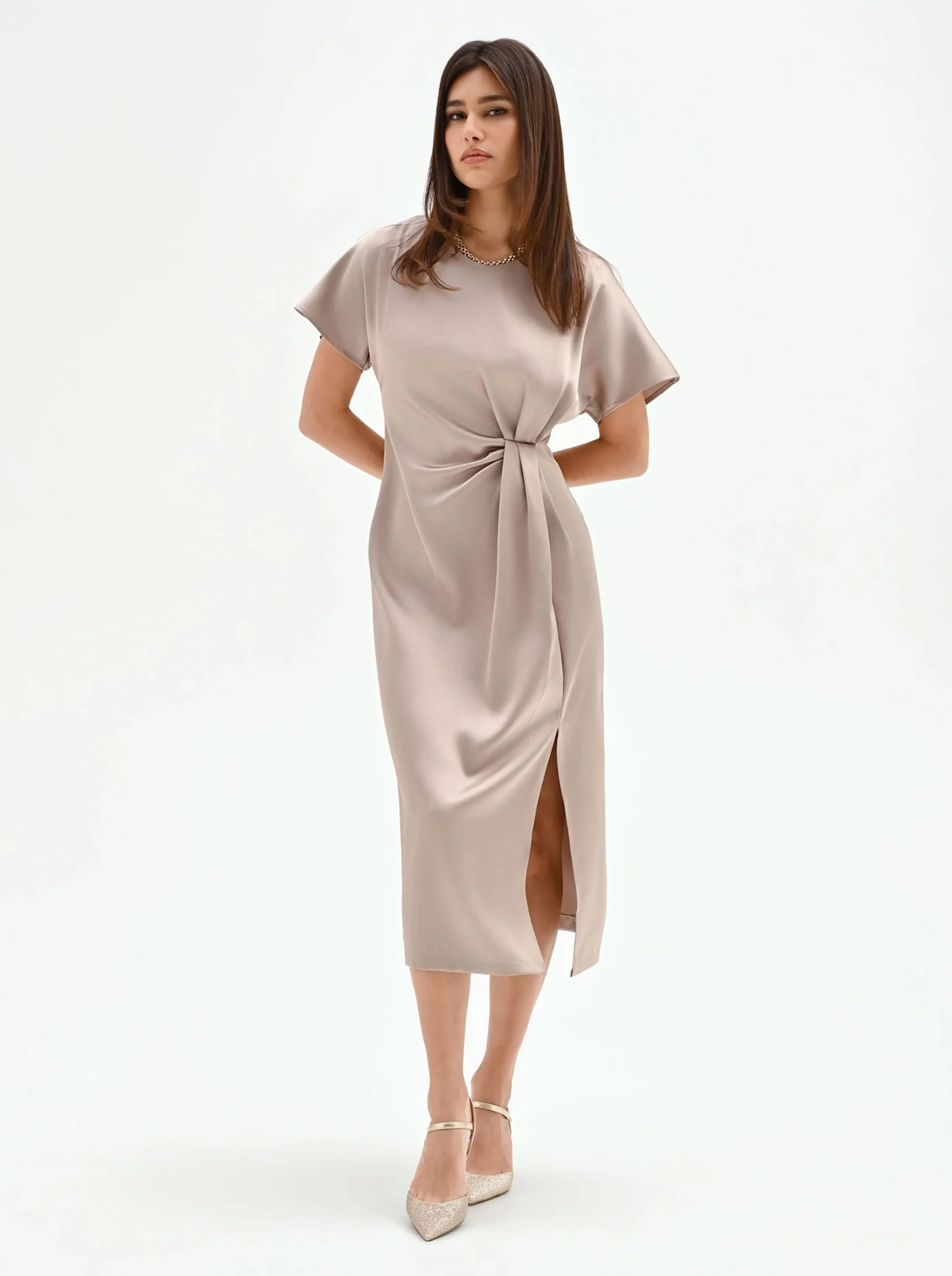 Woman in a taupe satin wrap dress with a side slit and short sleeves, stands confidently. She wears elegant heels, exuding a chic and poised vibe.