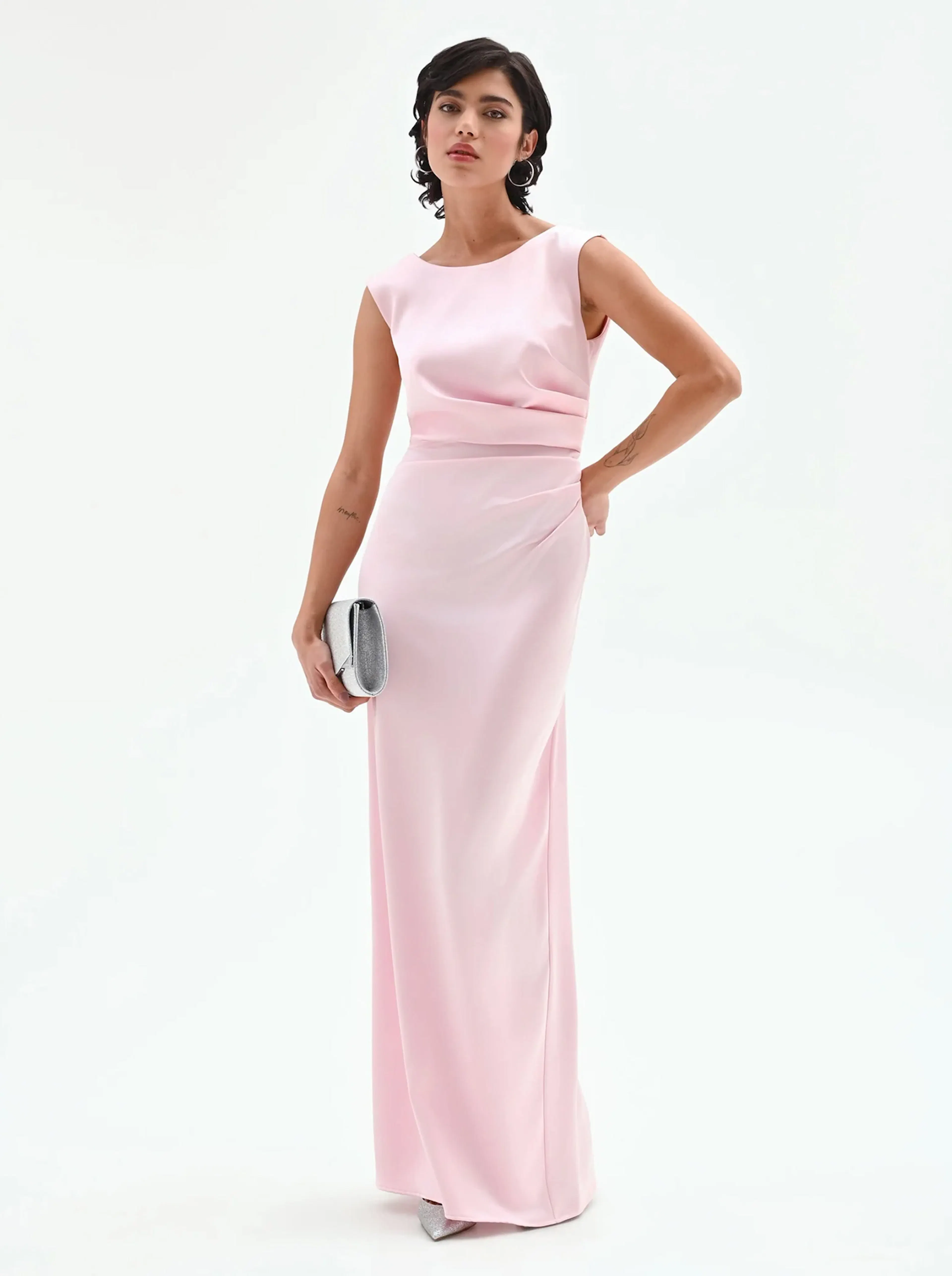 Woman in a sleeveless, floor-length pale pink gown holding a silver clutch purse, posing against a plain white background.