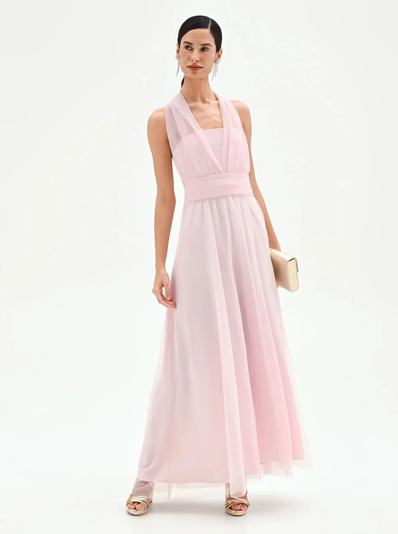 Woman wearing a sleeveless light pink floor-length dress with a sheer overlay, holding a beige clutch and wearing gold heeled sandals.