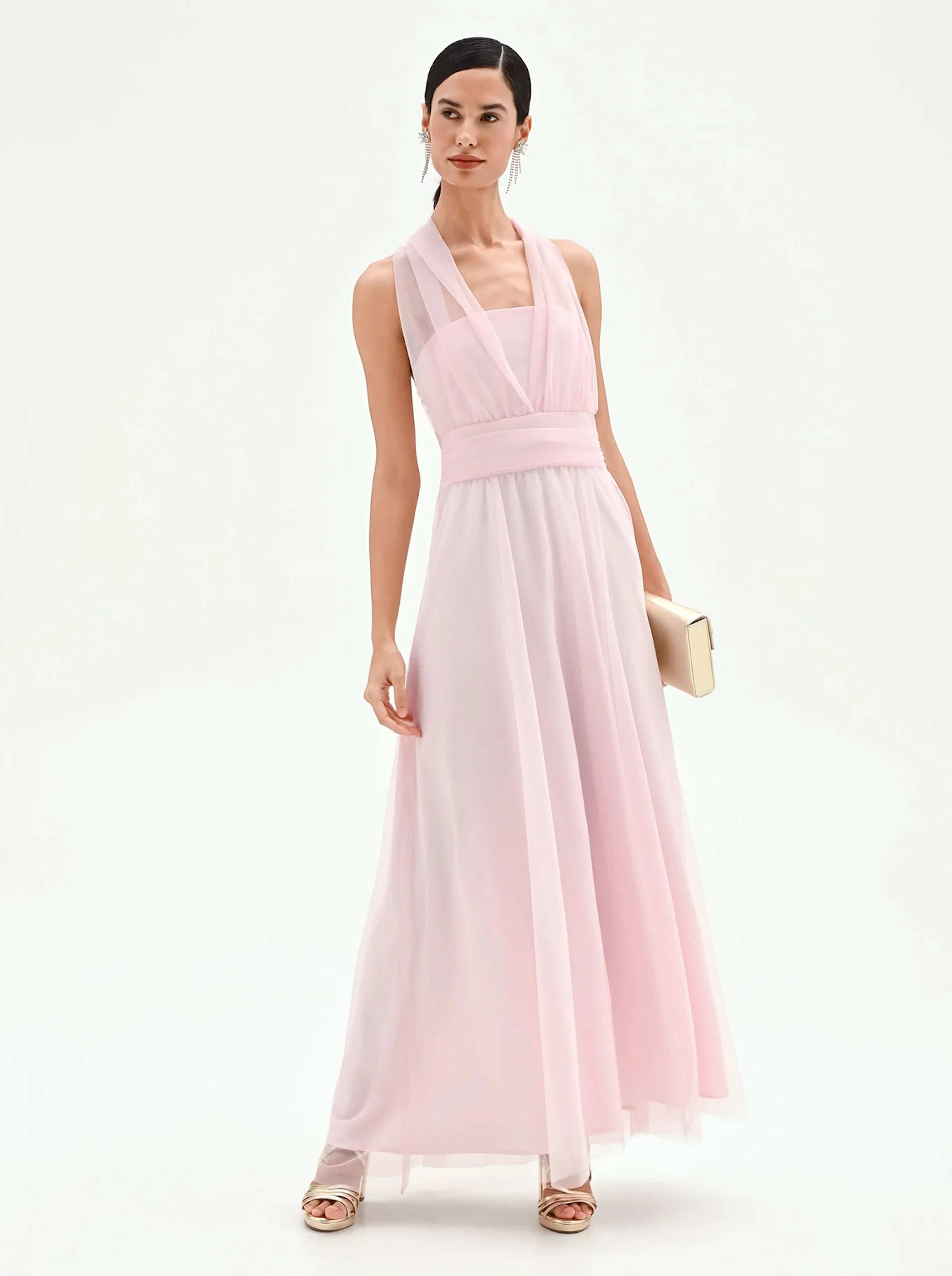 Woman wearing a sleeveless light pink floor-length dress with a sheer overlay, holding a beige clutch and wearing gold heeled sandals.