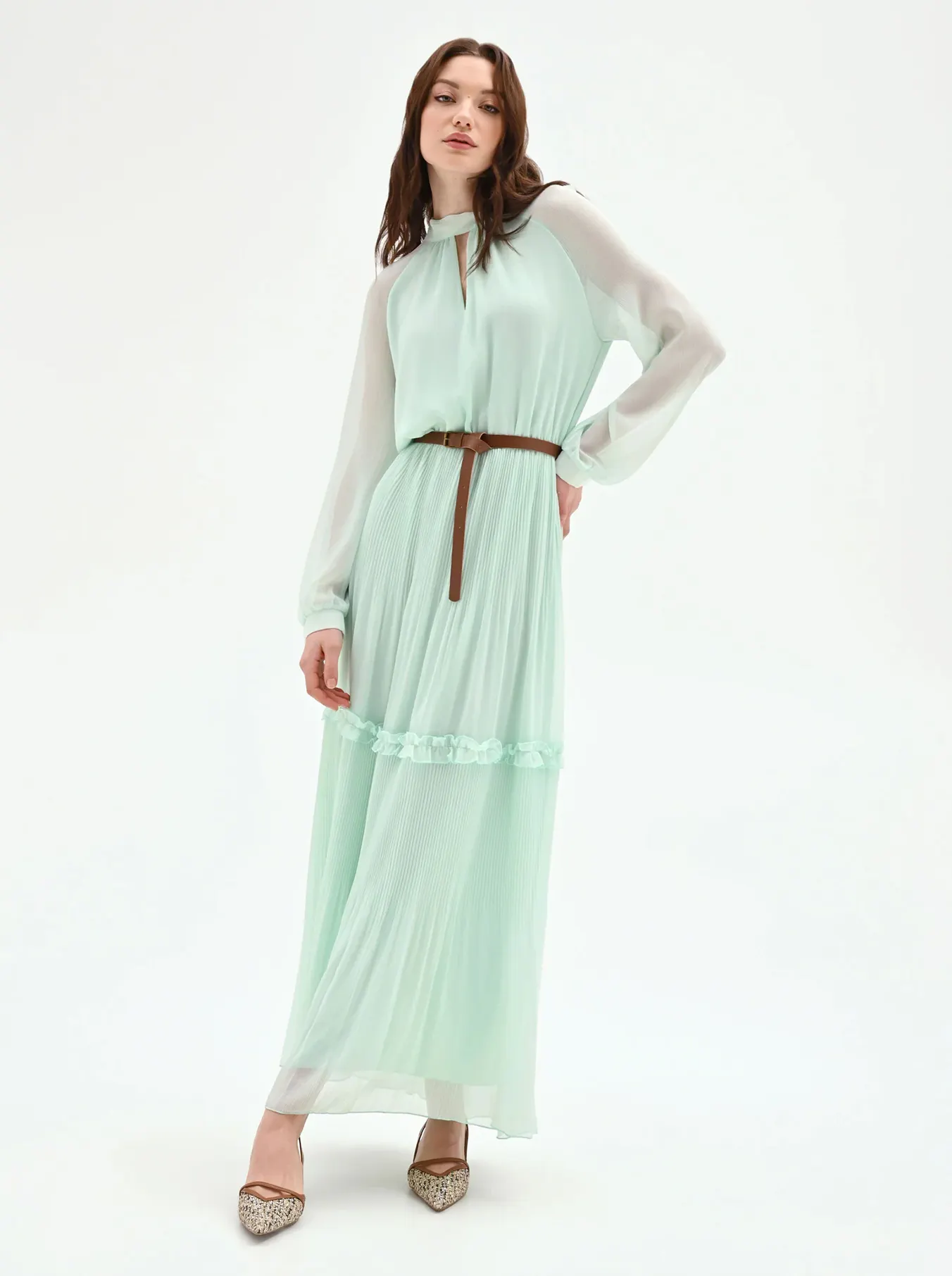 Woman wearing a mint green long-sleeve pleated maxi dress with a brown belt and patterned pointed-toe heels, standing against a white background.