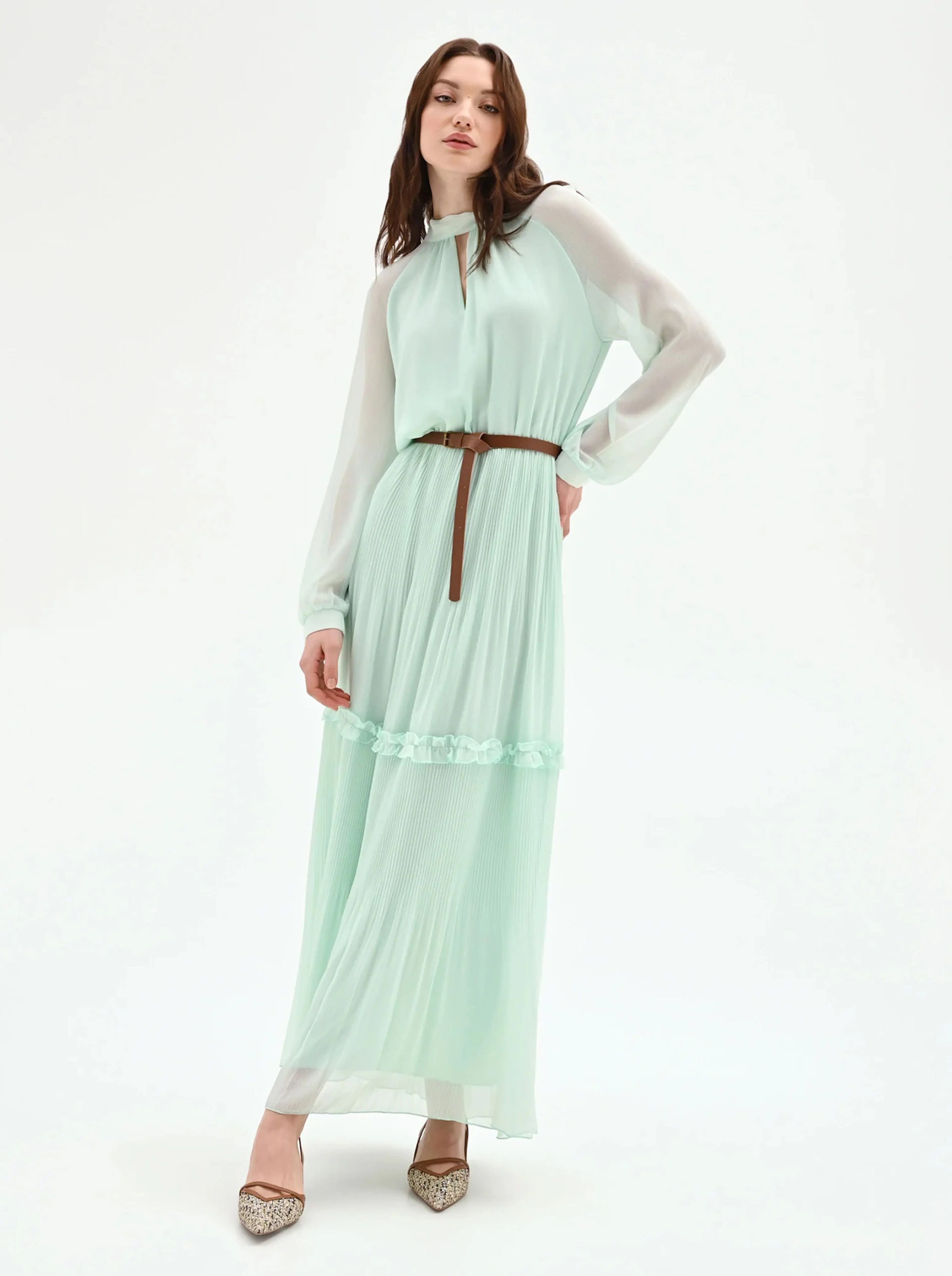 Woman wearing a mint green long-sleeve pleated maxi dress with a brown belt and patterned pointed-toe heels, standing against a white background.