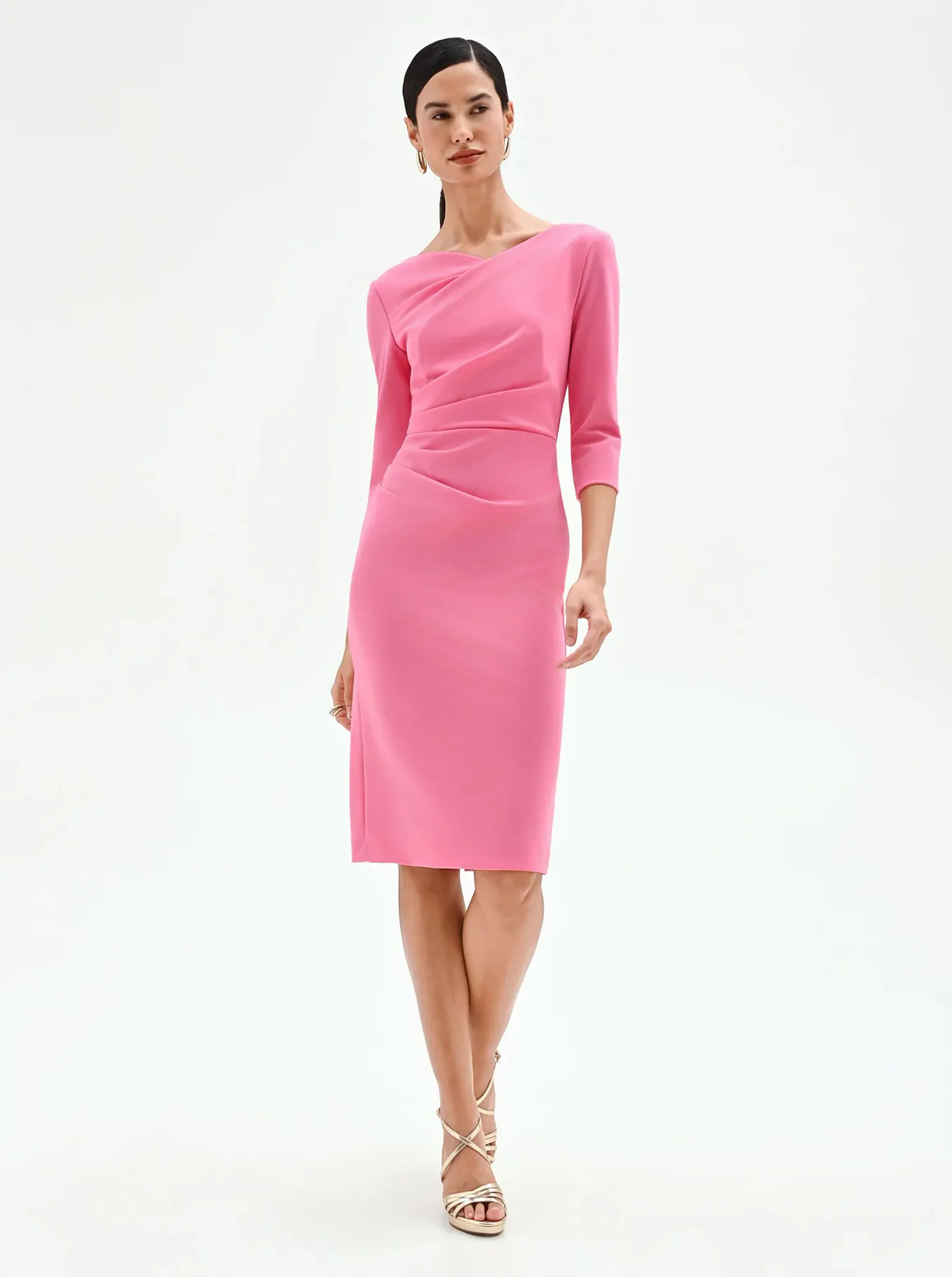 A woman in a pink, knee-length, form-fitting dress with three-quarter sleeves stands gracefully. She wears strappy heels, exuding elegance against a white background.
