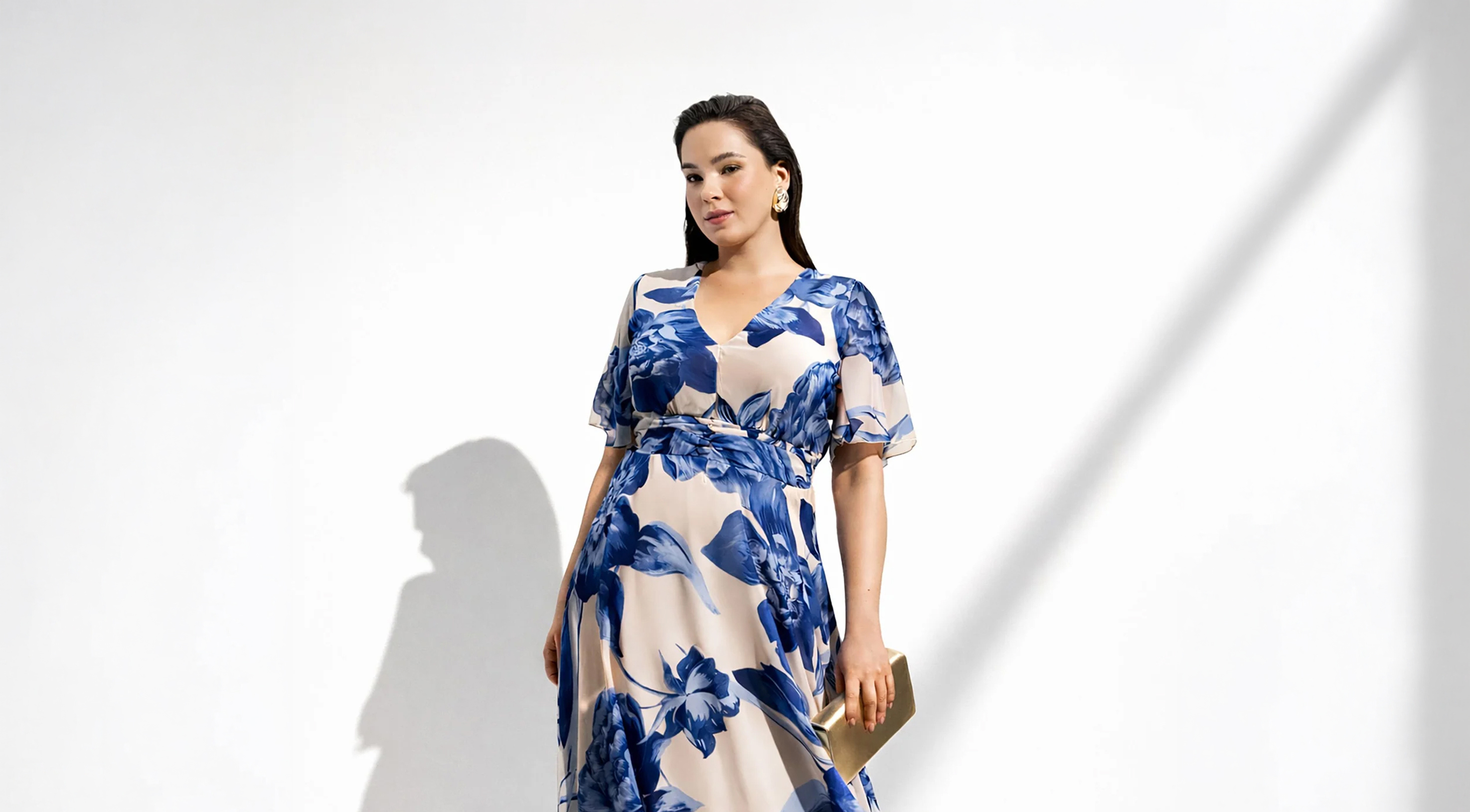 Woman in a blue and white floral dress holding a gold clutch against a plain white background with shadow lines.