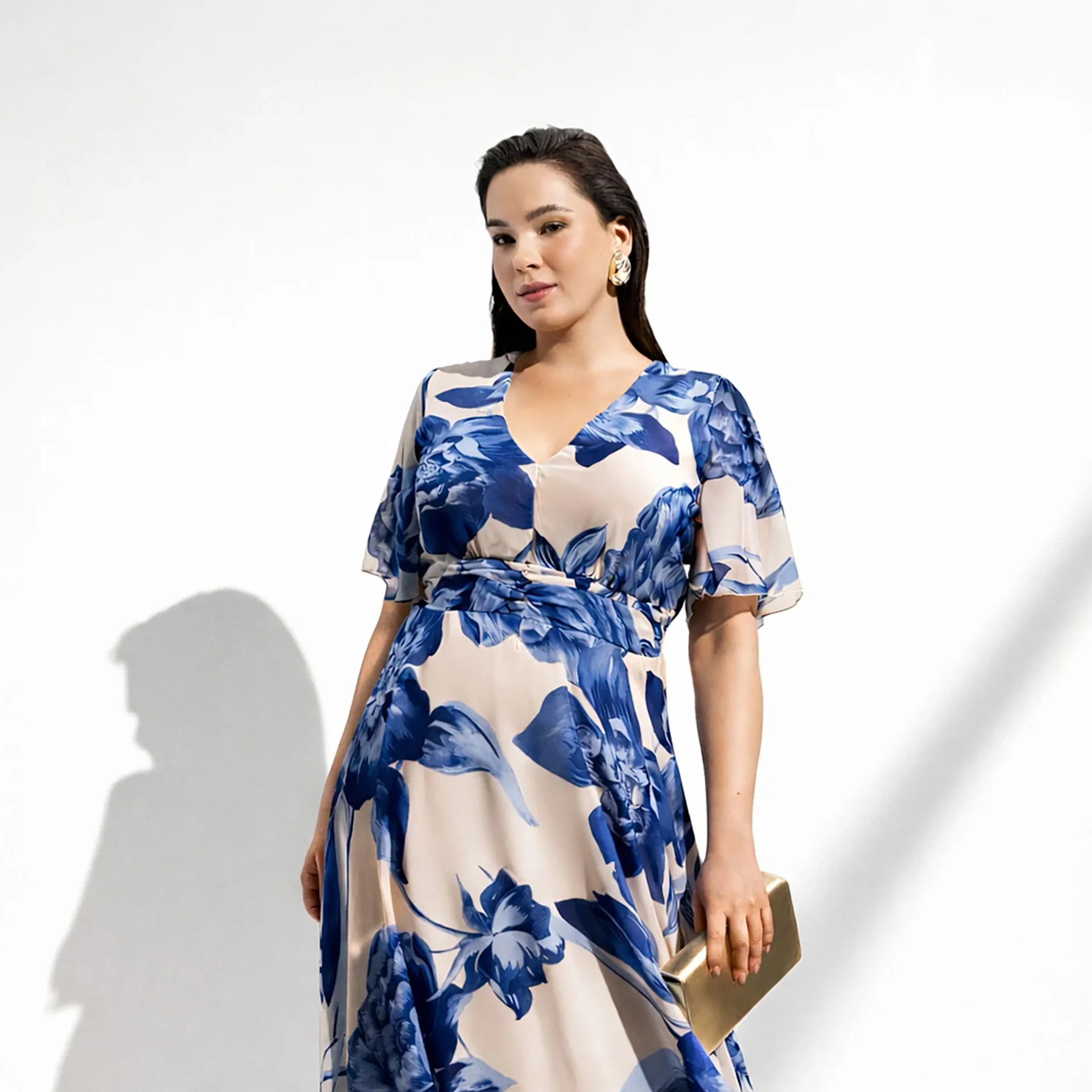 Woman in a blue and white floral dress holding a gold clutch against a plain white background with shadow lines.
