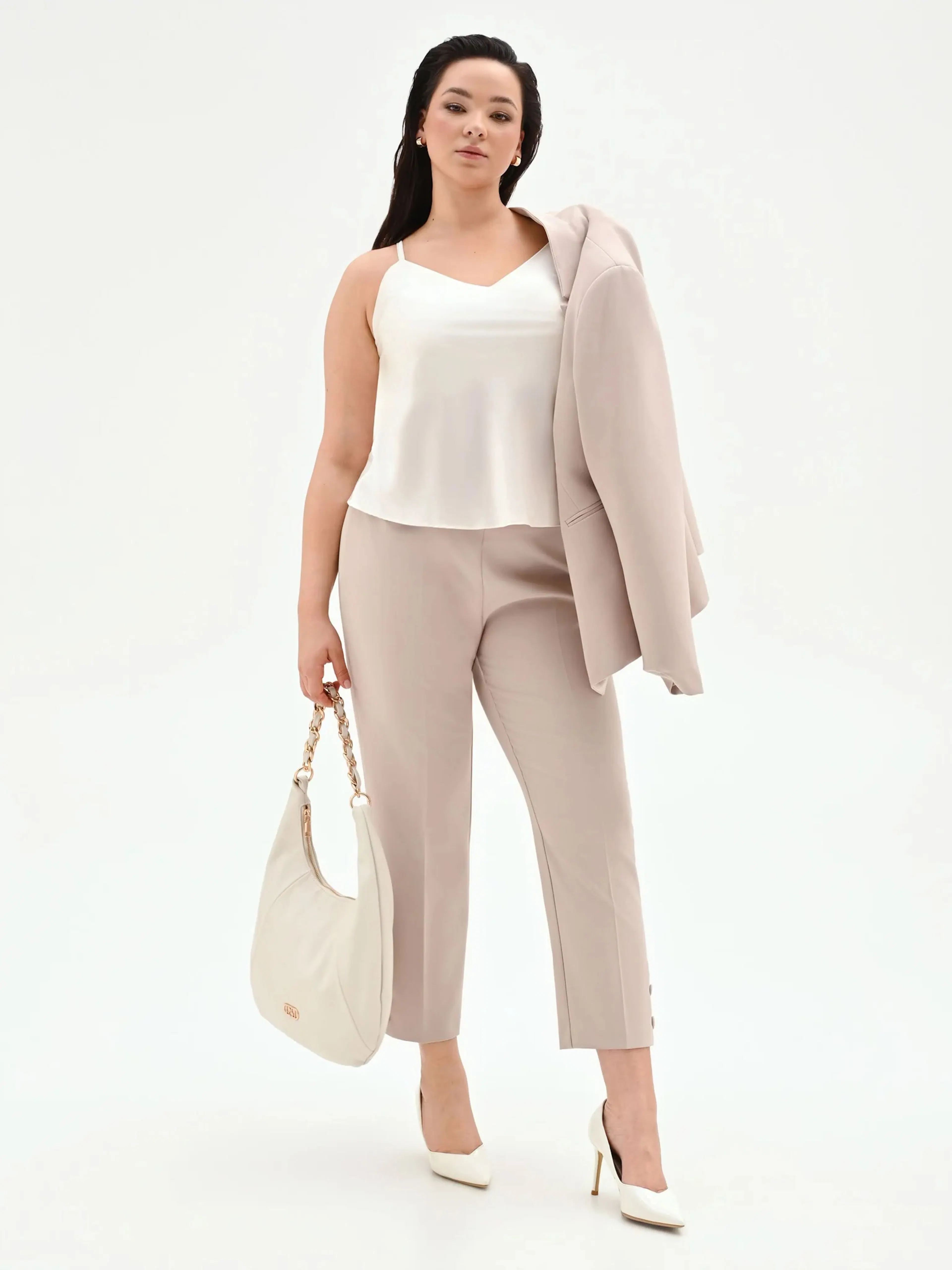 Woman in a stylish cream outfit holds a matching blazer over her shoulder. She carries a white handbag and wears white heels, exuding confidence and elegance.