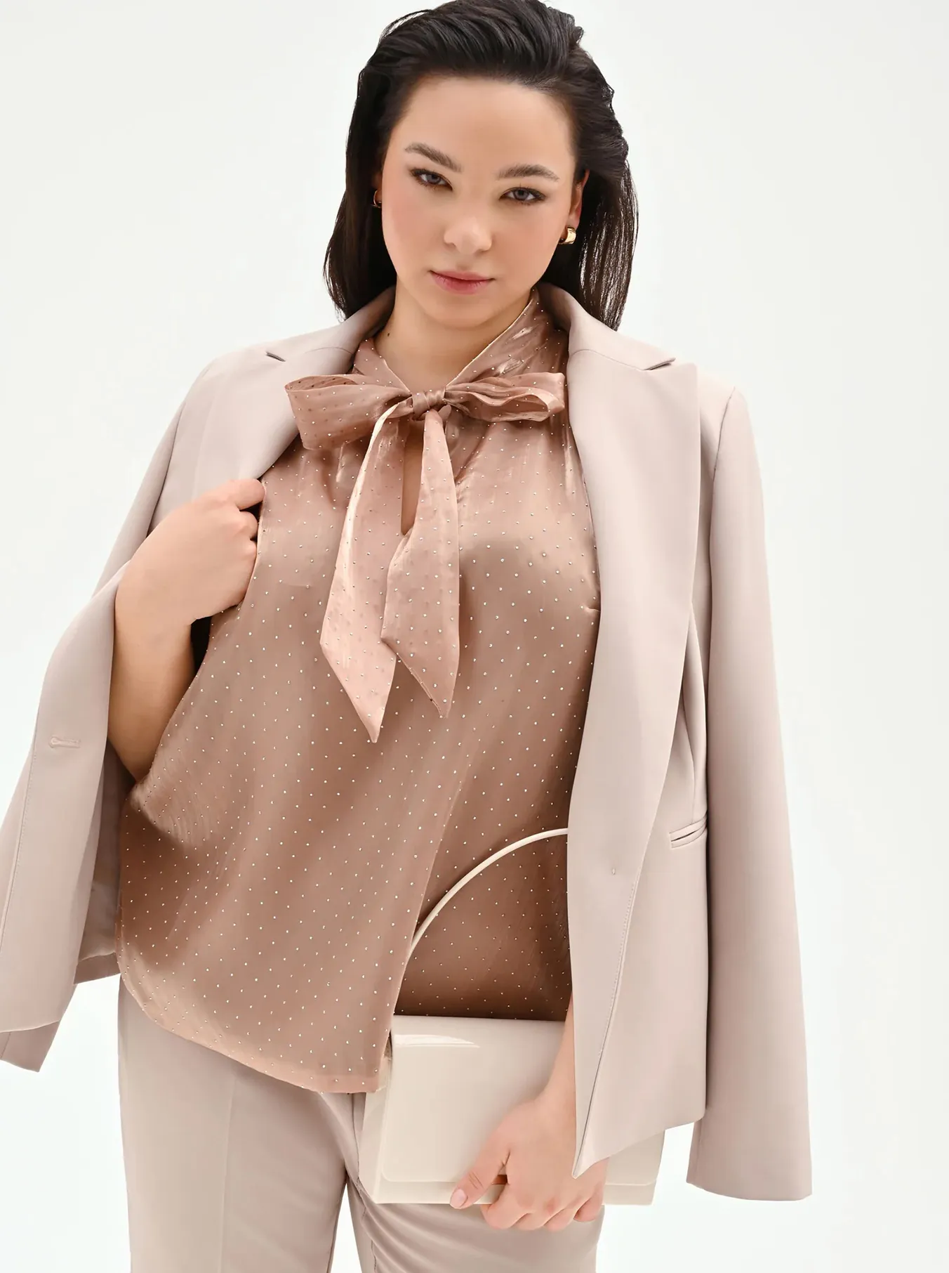 Person wearing a beige suit draped over shoulders with a light brown polka-dot bow blouse, holding a white handbag against a plain background.