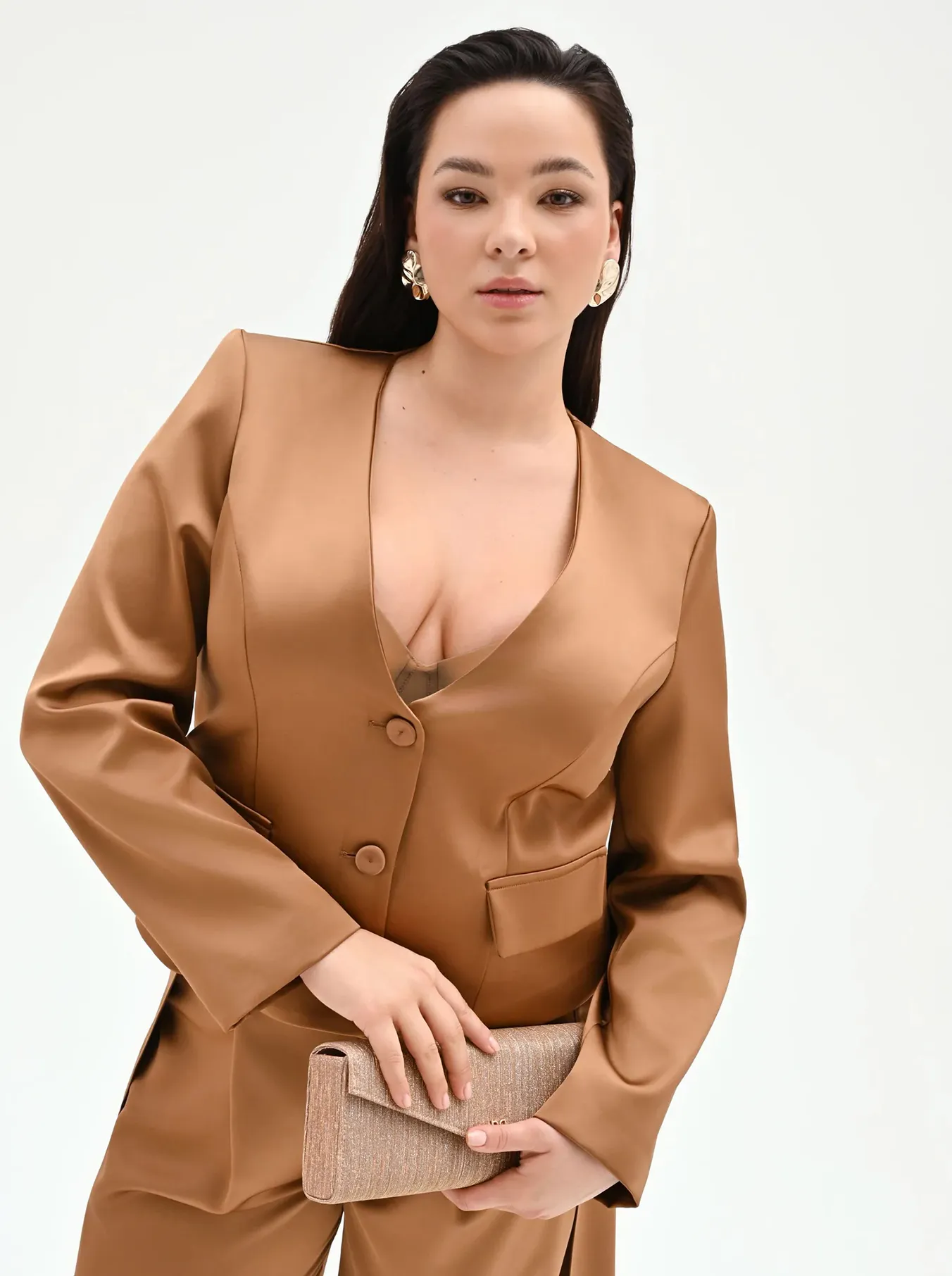 Woman in a tan satin blazer and pants holding a textured beige clutch against a plain white background.