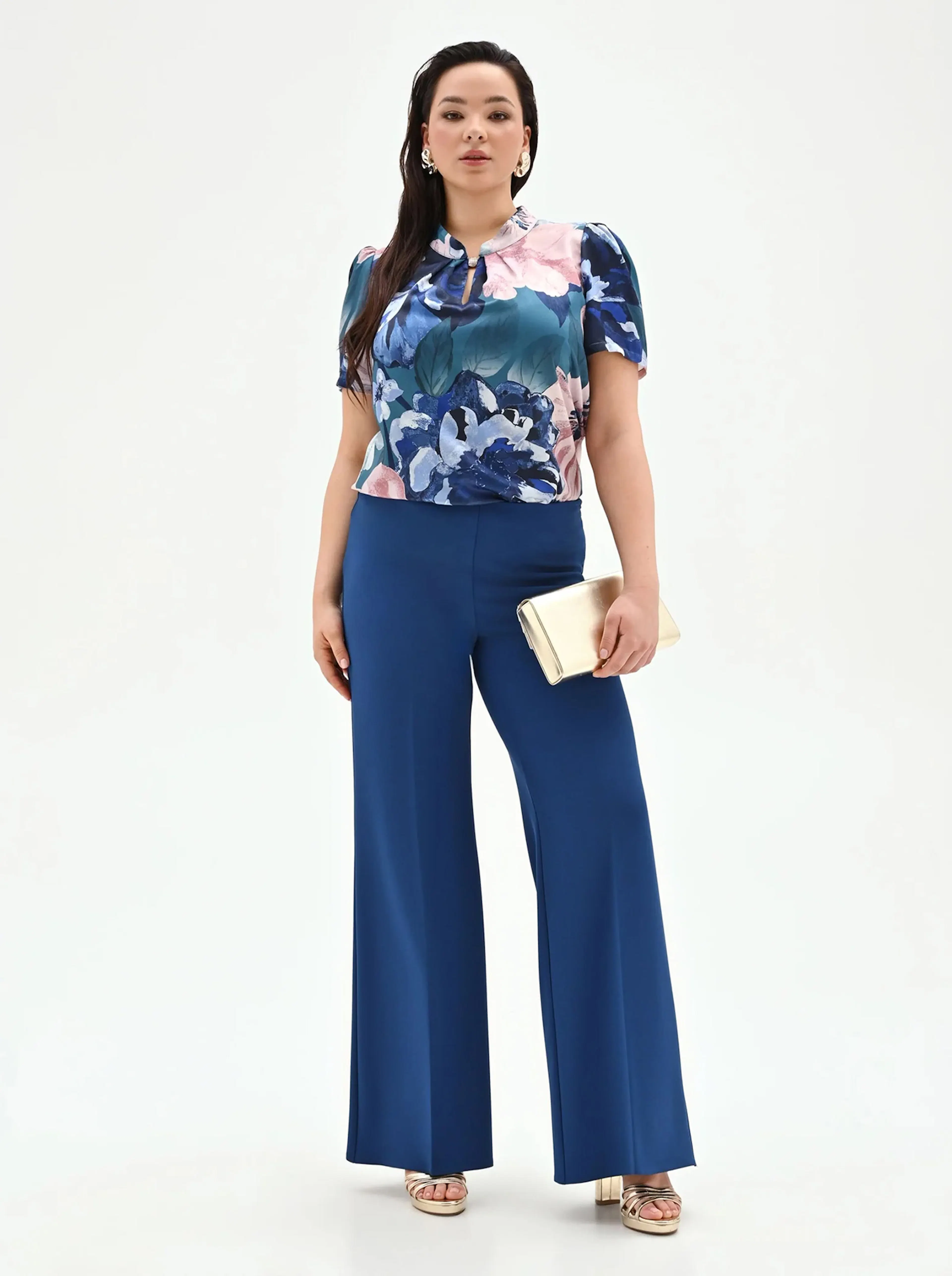 Woman wearing a floral blue and pink blouse paired with high-waisted wide-leg blue pants, gold sandals, and holding a gold clutch.