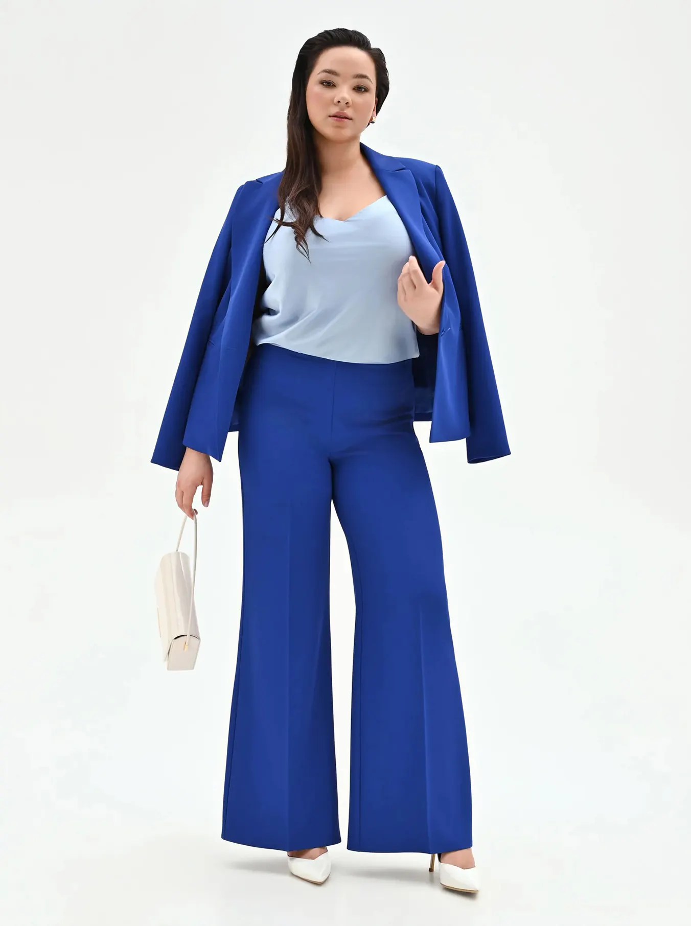 Woman wearing a bright blue pantsuit with a light blue top, white heels, and holding a small white handbag against a plain background.