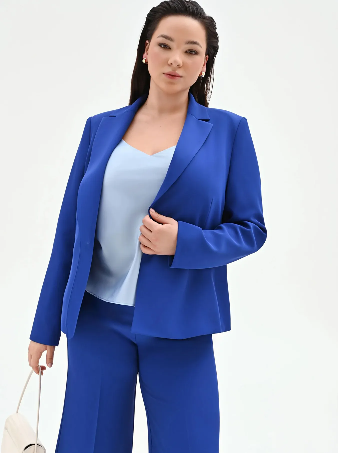 Woman in a bright blue suit with wide-leg pants and light blue top, holding a white handbag against a plain white background.