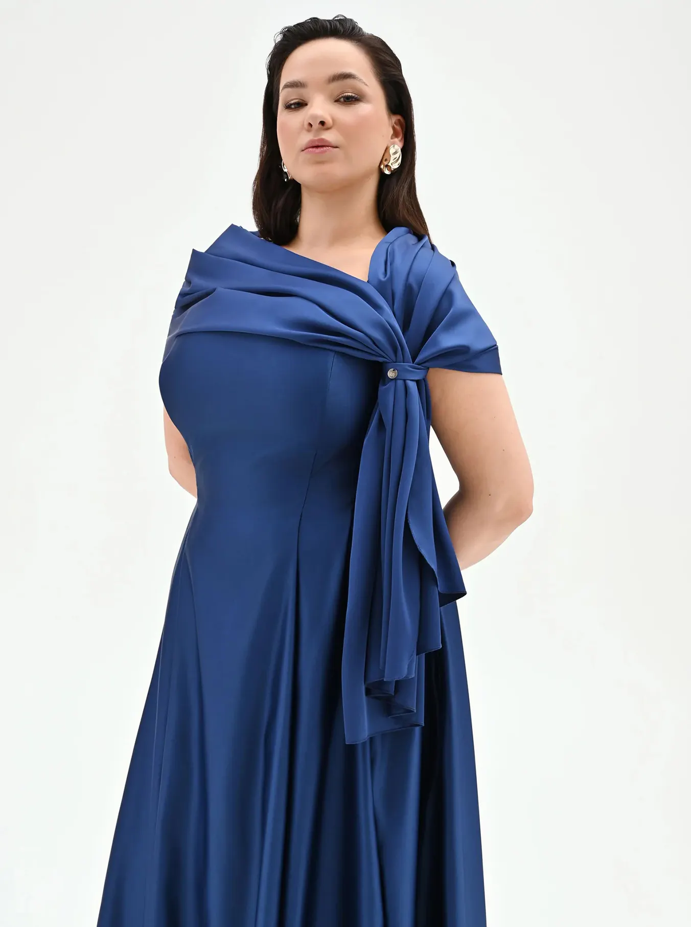 Woman in an elegant blue dress with a flowing, draped design, standing confidently against a white background. She wears bold earrings, exuding poise.