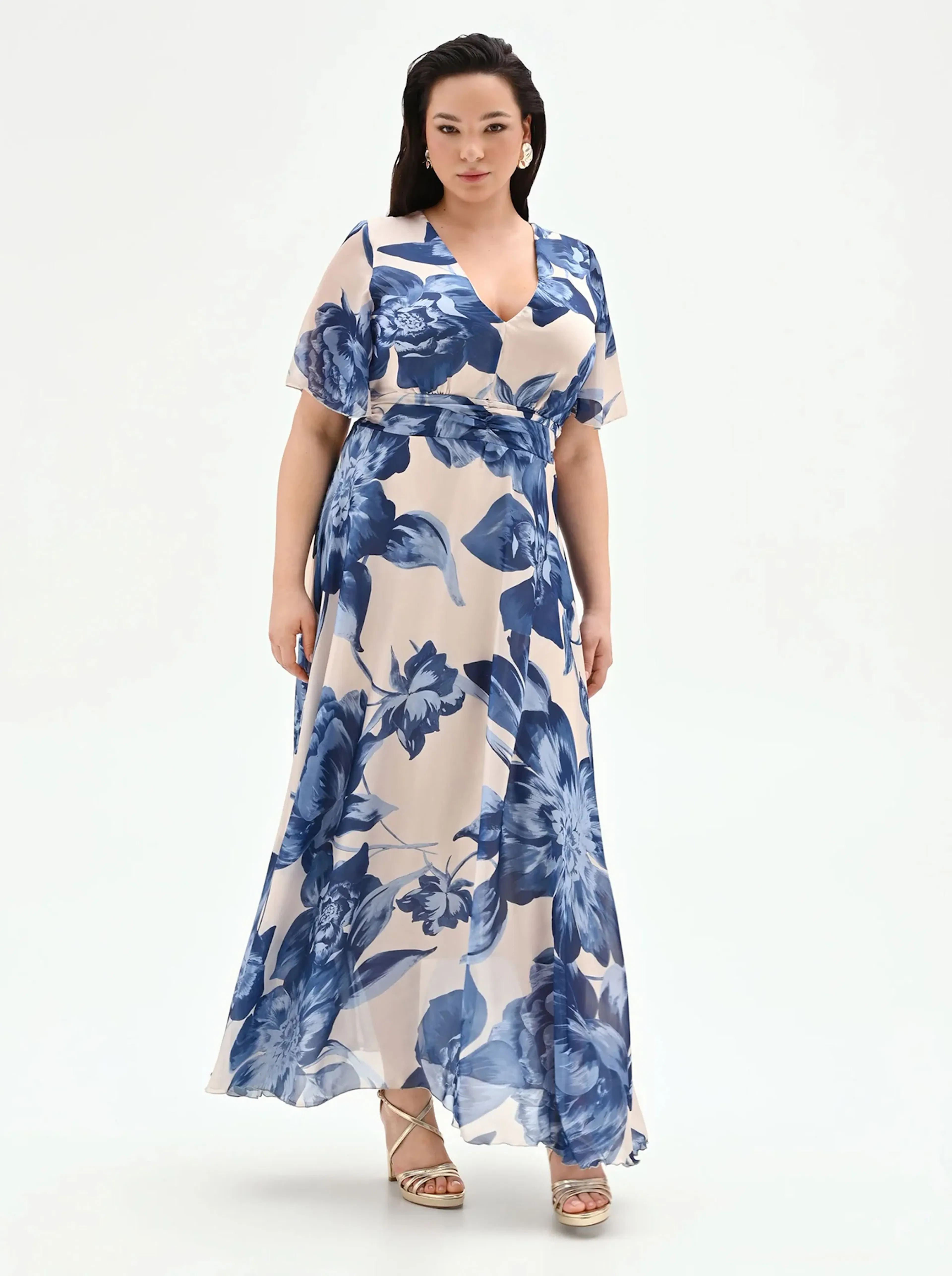 Woman in a flowing blue floral dress stands confidently against a white background, holding a gold clutch. Elegant and poised, conveying a chic, summery vibe.