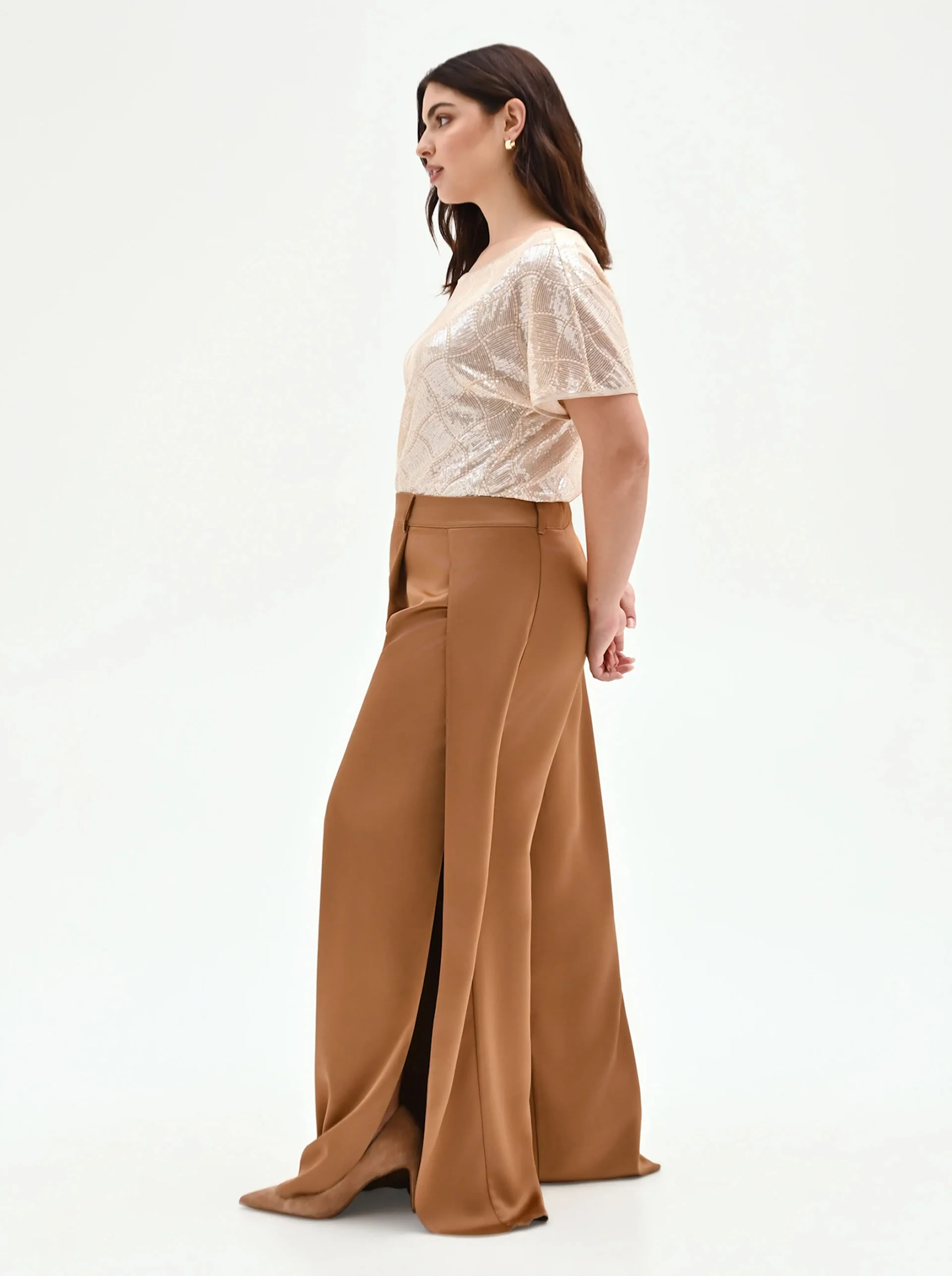 Woman in elegant outfit: beige patterned top, brown wide-leg trousers. She holds a small purse, posing confidently against a plain backdrop.
