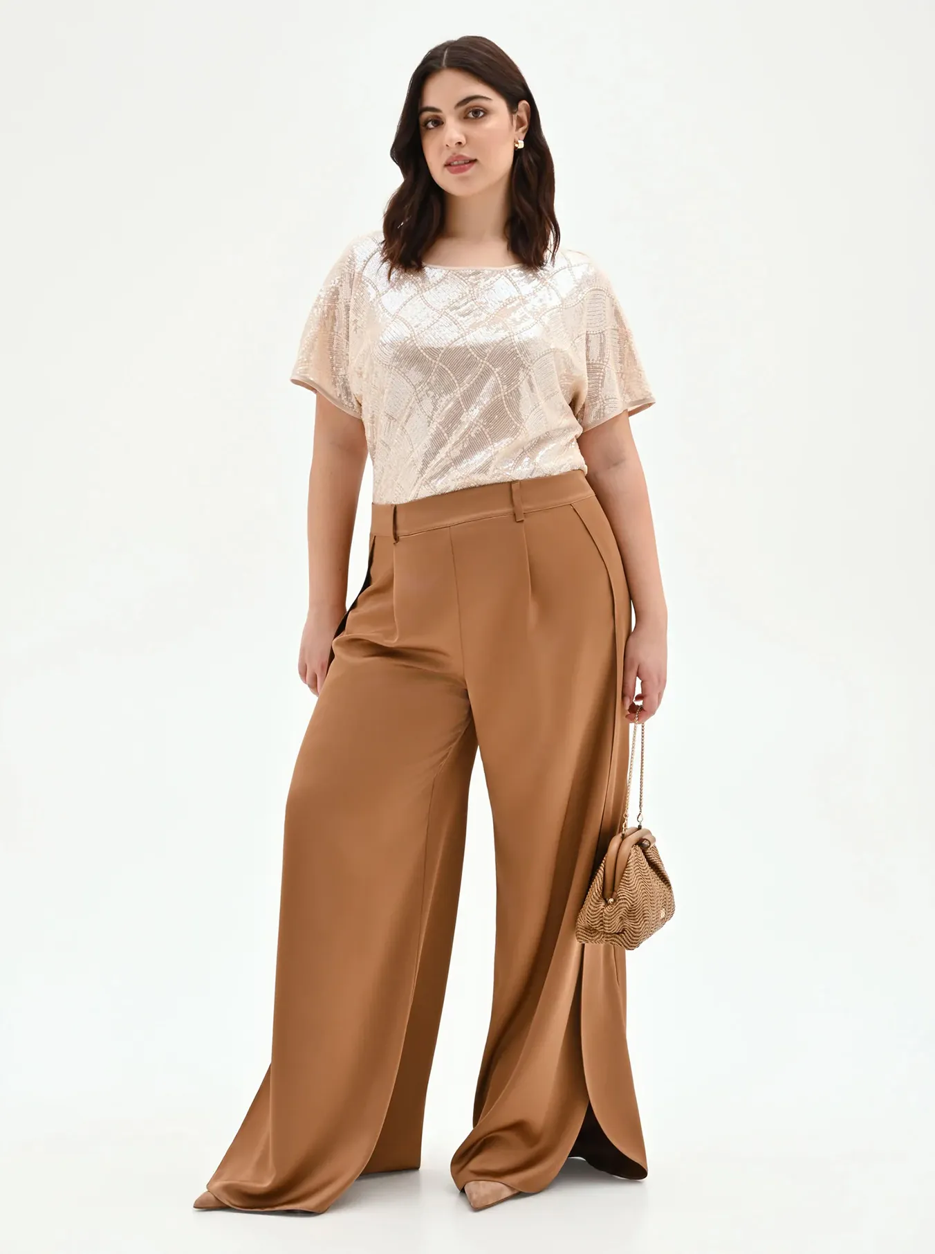 Woman in elegant outfit: beige patterned top, brown wide-leg trousers. She holds a small purse, posing confidently against a plain backdrop.