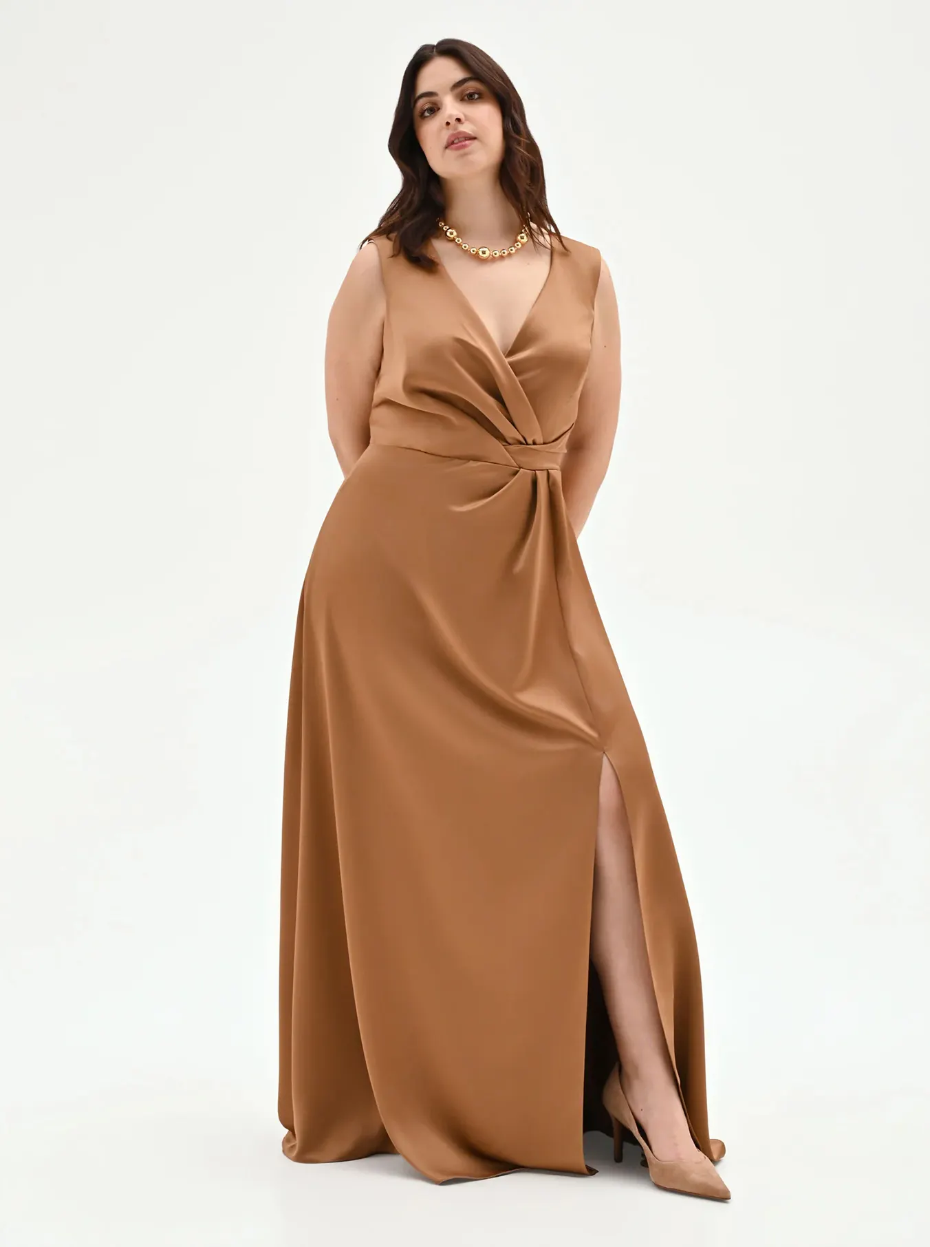 Woman in a sleeveless bronze gown with a deep V-neck and high slit, holding a clutch. She wears a gold necklace and nude heels, exuding elegance.