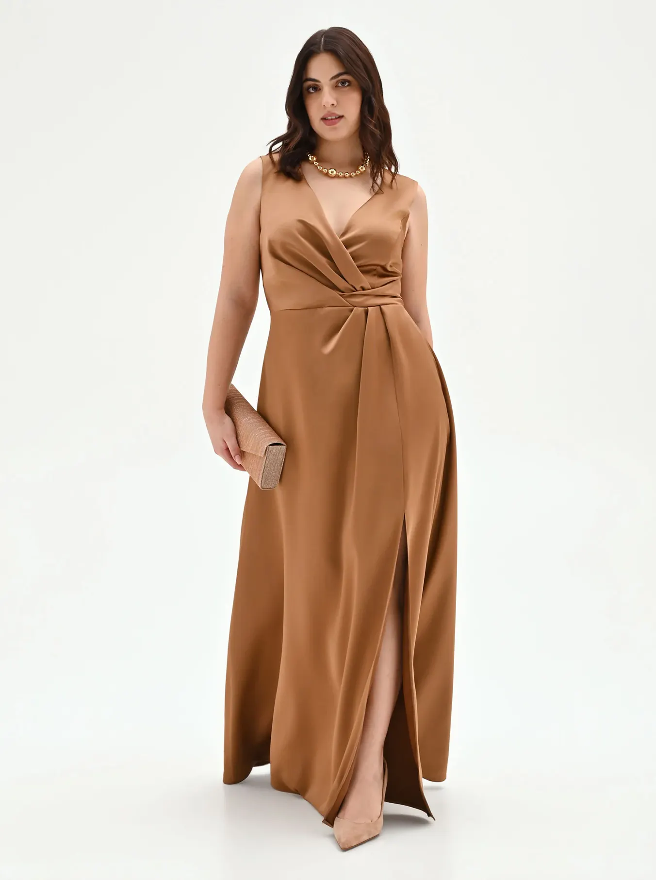 Woman in a sleeveless bronze gown with a deep V-neck and high slit, holding a clutch. She wears a gold necklace and nude heels, exuding elegance.