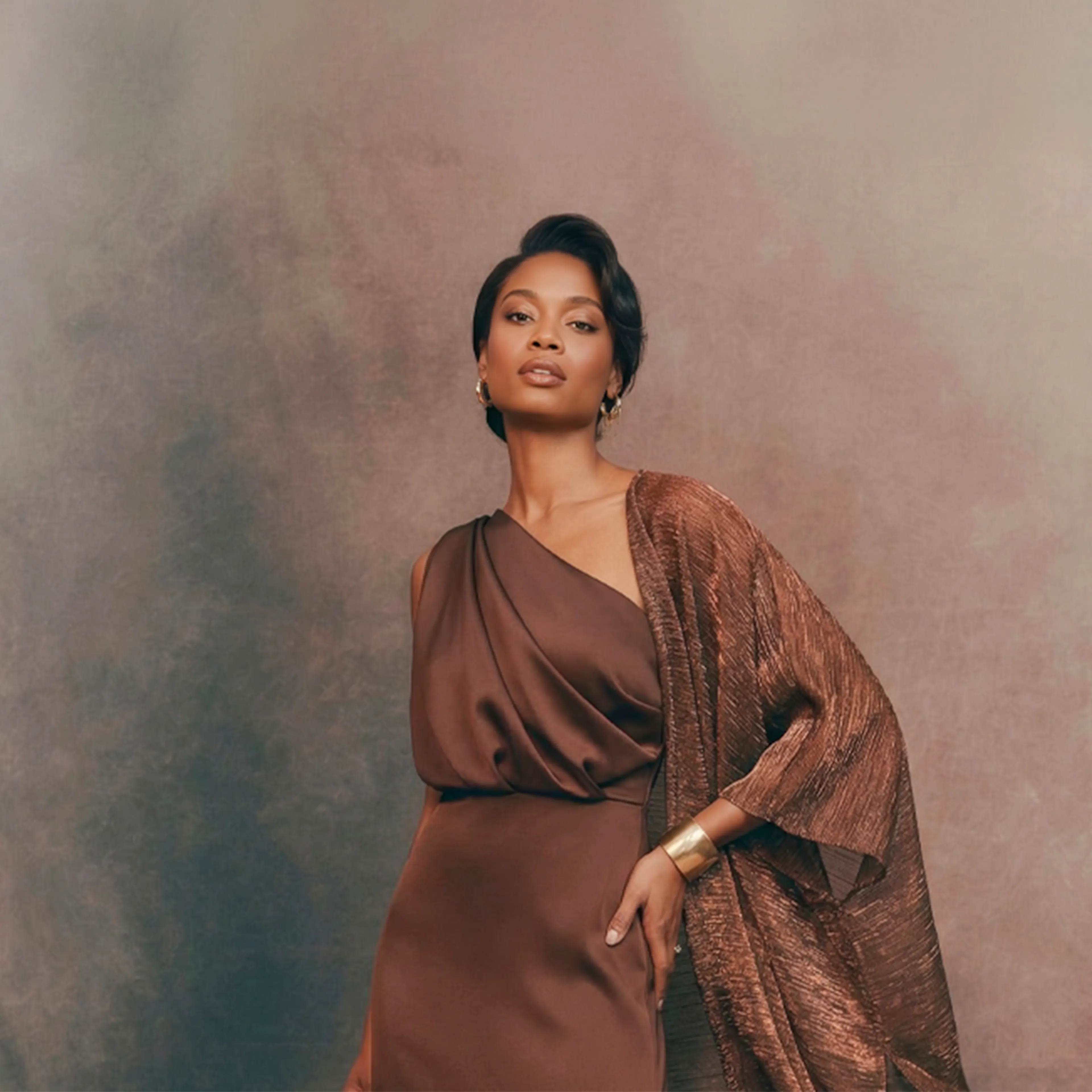 A woman in an elegant brown dress and shawl stands confidently against a soft, neutral-toned backdrop. Her posture conveys grace and poise.