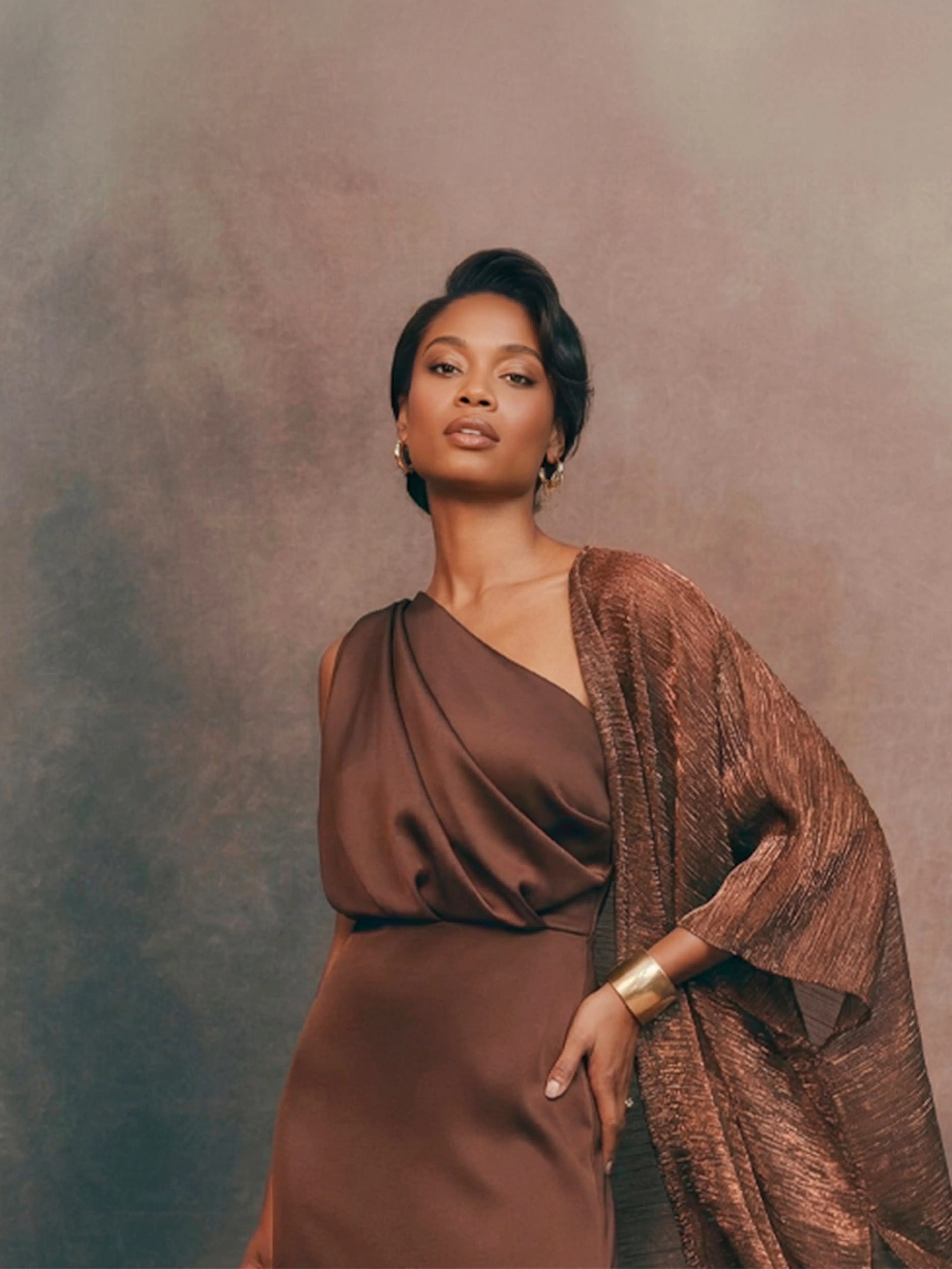 A woman in an elegant brown dress and shawl stands confidently against a soft, neutral-toned backdrop. Her posture conveys grace and poise.