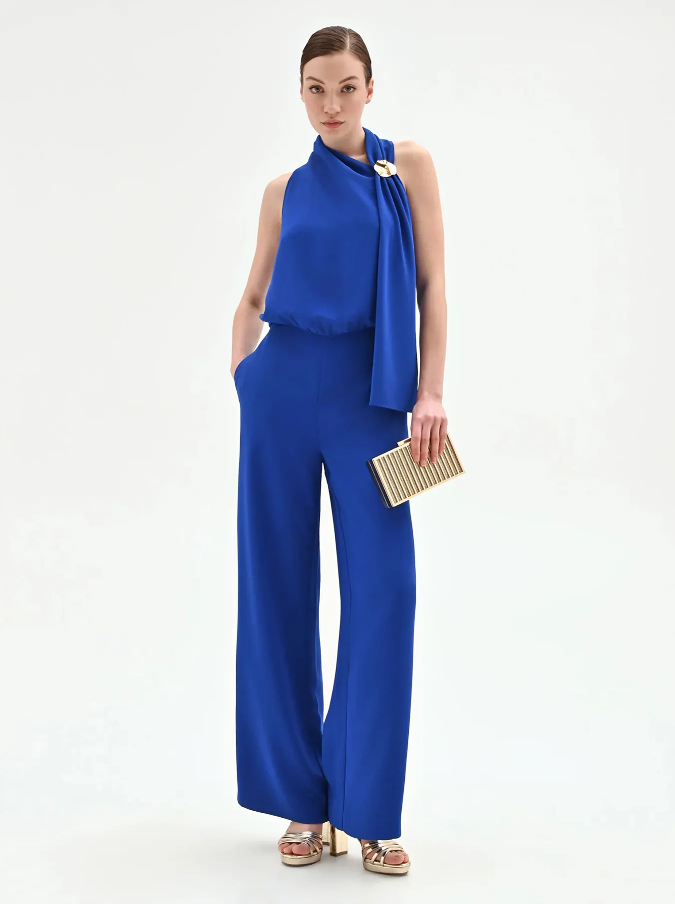 Woman in elegant blue jumpsuit with a gold clasp and high neckline, holding a gold clutch. She wears gold platform sandals, exuding sophistication.