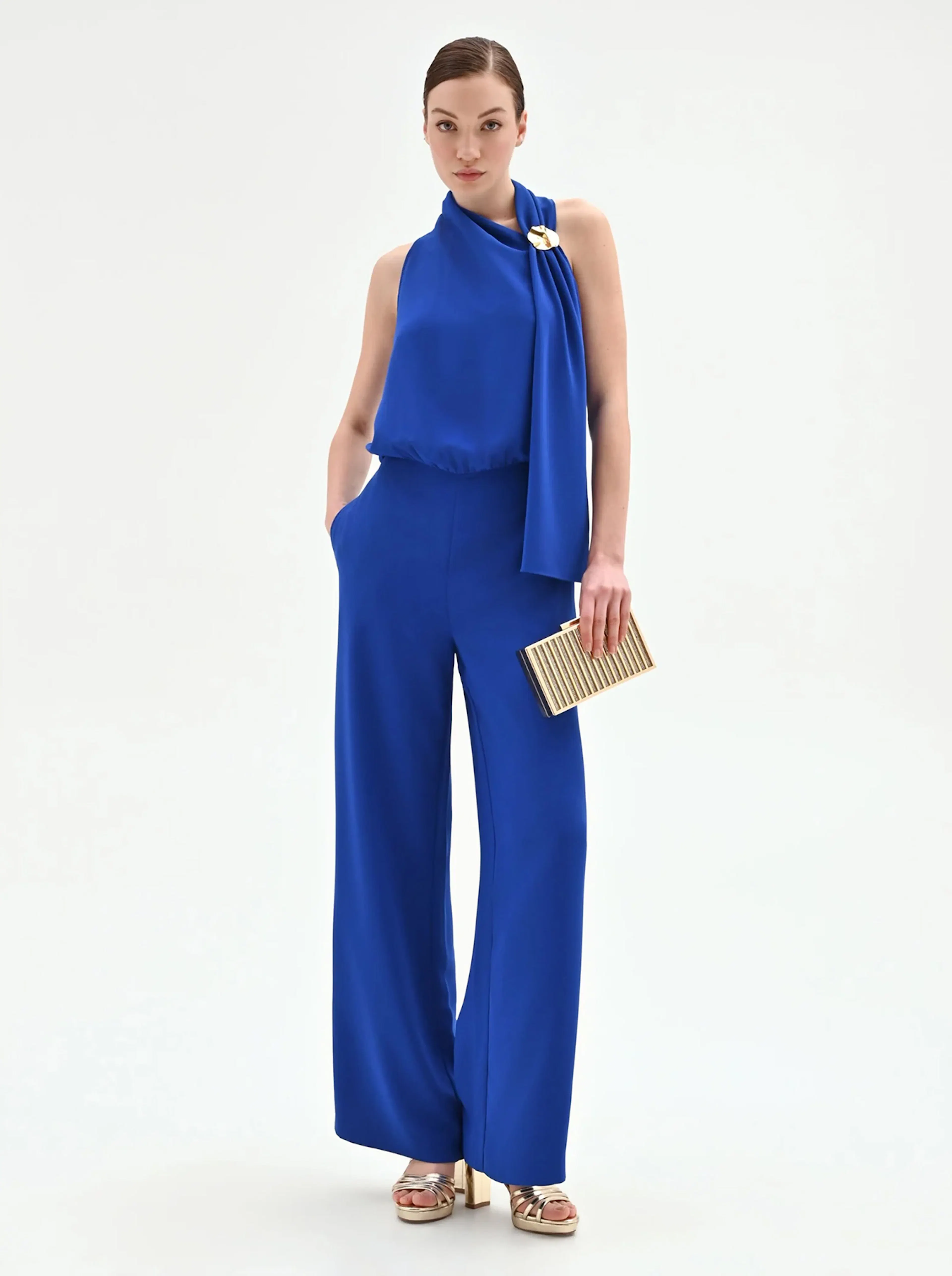 Woman in elegant blue jumpsuit with a gold clasp and high neckline, holding a gold clutch. She wears gold platform sandals, exuding sophistication.