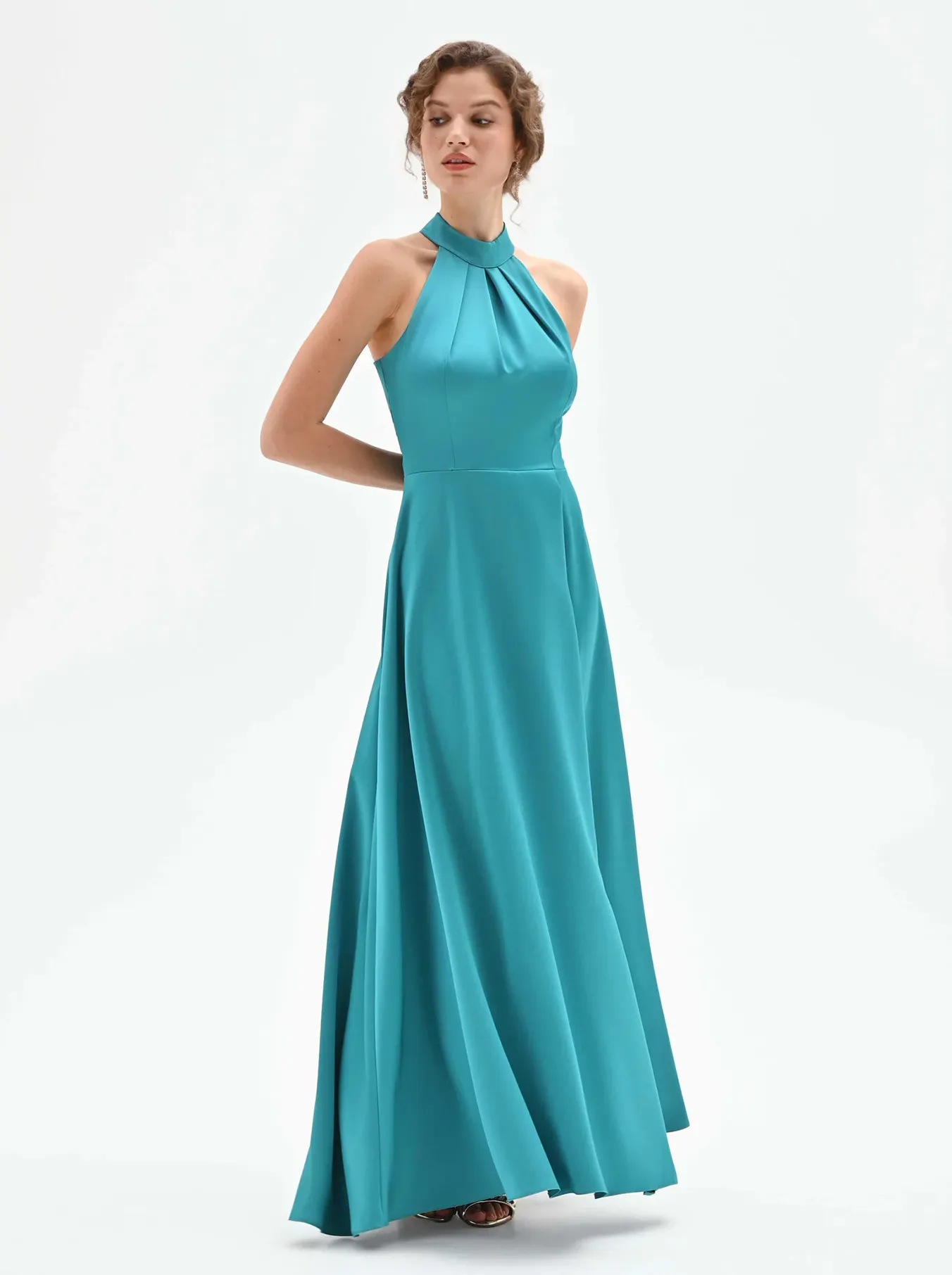 A woman in a turquoise halter-neck gown stands with hands behind her back. The dress flows elegantly, conveying a serene and sophisticated tone.