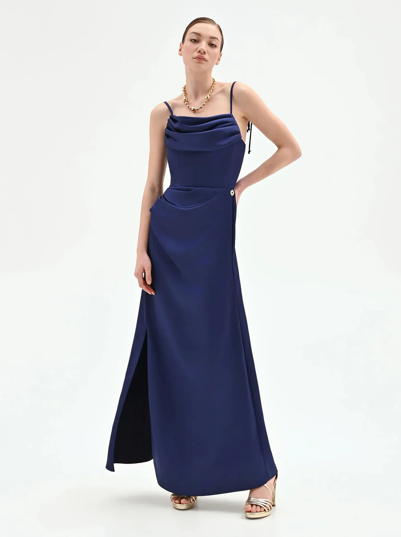 A woman in a navy blue gown with thin straps and a side slit poses confidently. She wears a gold necklace and high heels, exuding elegance and poise.