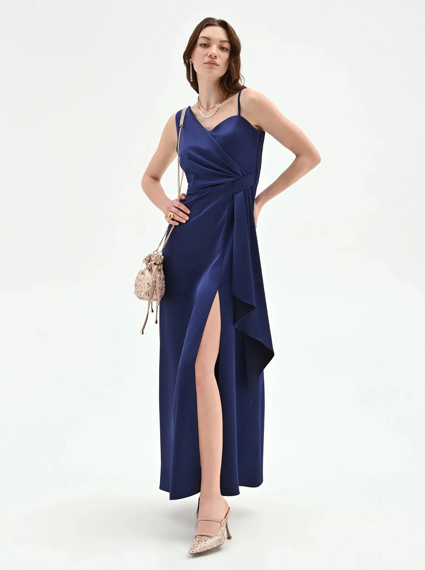 Woman in a sleek blue satin gown with a high slit poses confidently, wearing beige heels and holding a small sparkling handbag, exuding elegance.