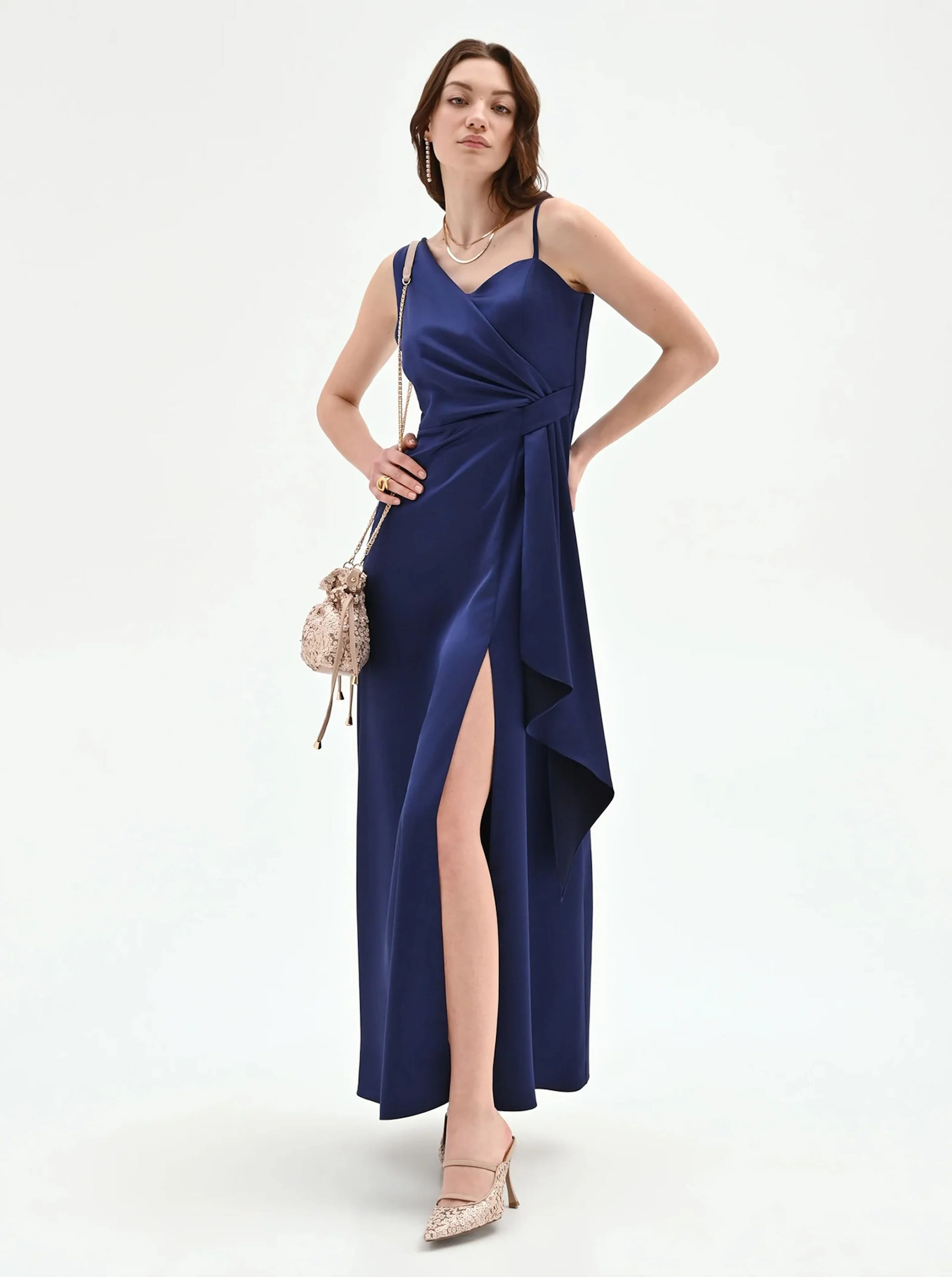 Woman in a sleek blue satin gown with a high slit poses confidently, wearing beige heels and holding a small sparkling handbag, exuding elegance.