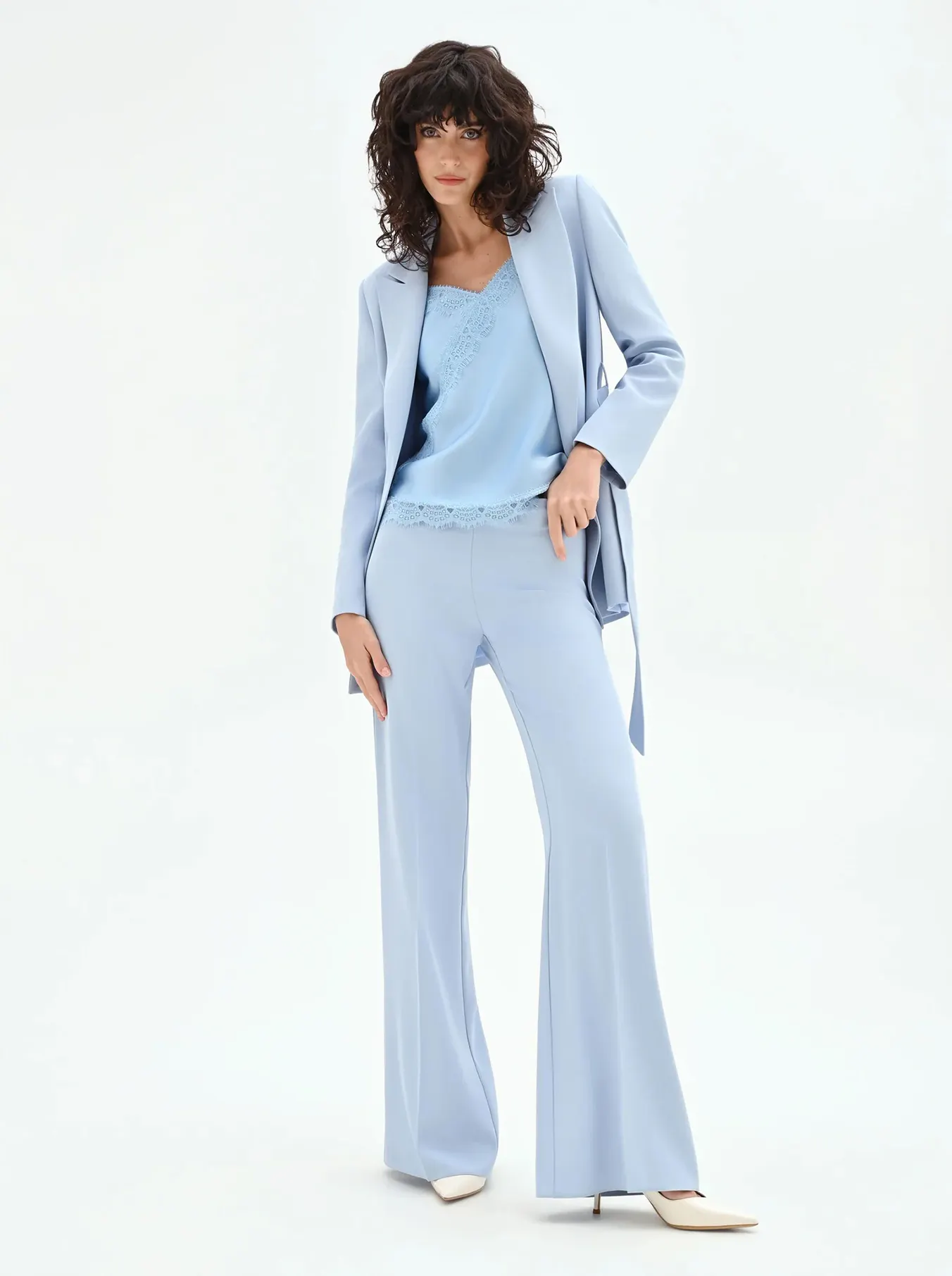 A woman with curly hair wears a light blue pantsuit with a matching camisole, striking a confident pose. The tone is elegant and modern.