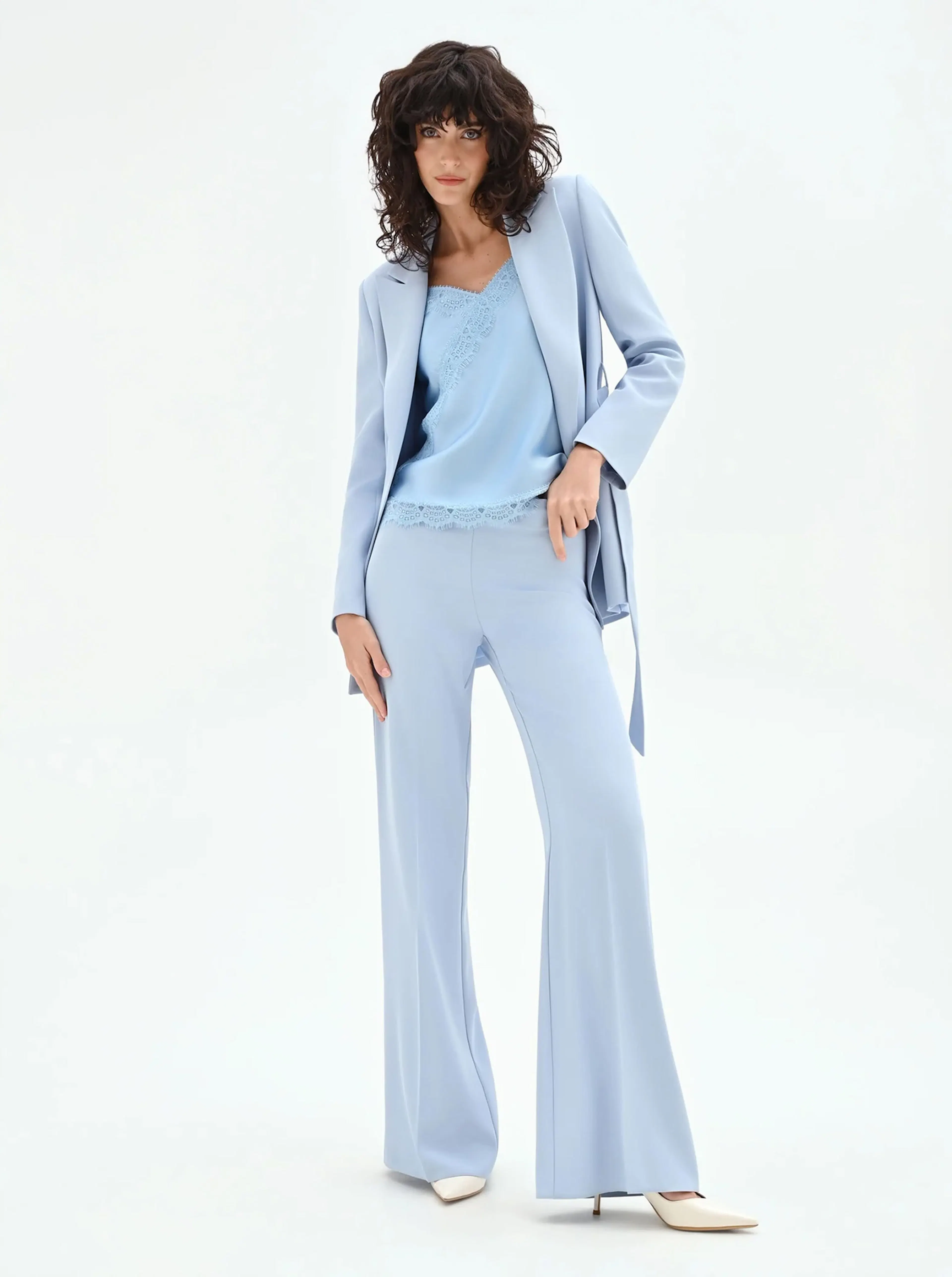 A woman with curly hair wears a light blue pantsuit with a matching camisole, striking a confident pose. The tone is elegant and modern.