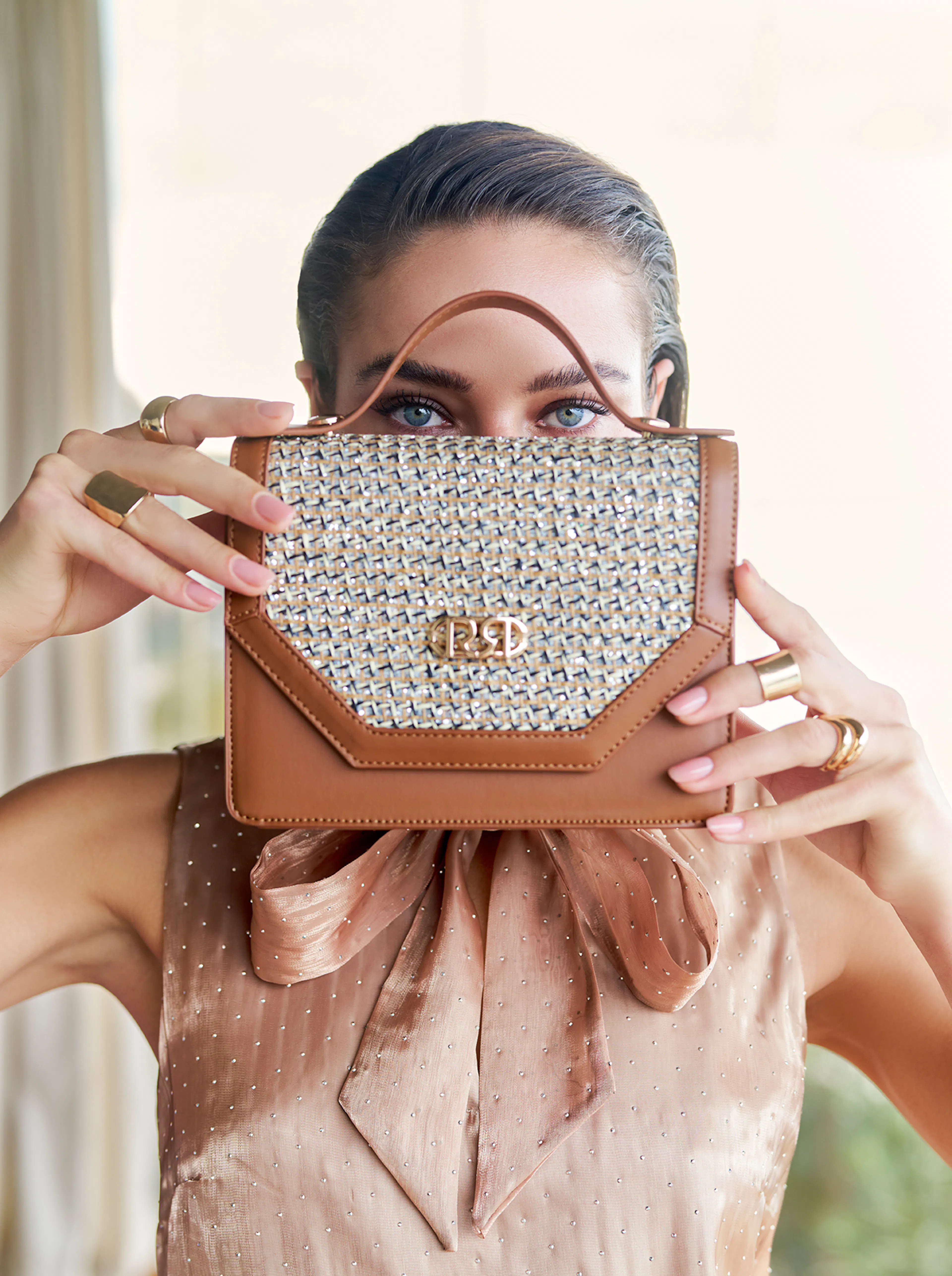 A person holds a stylish brown and woven textured handbag up to their face, revealing only their eyes. They wear a satin blouse and gold rings, conveying elegance.