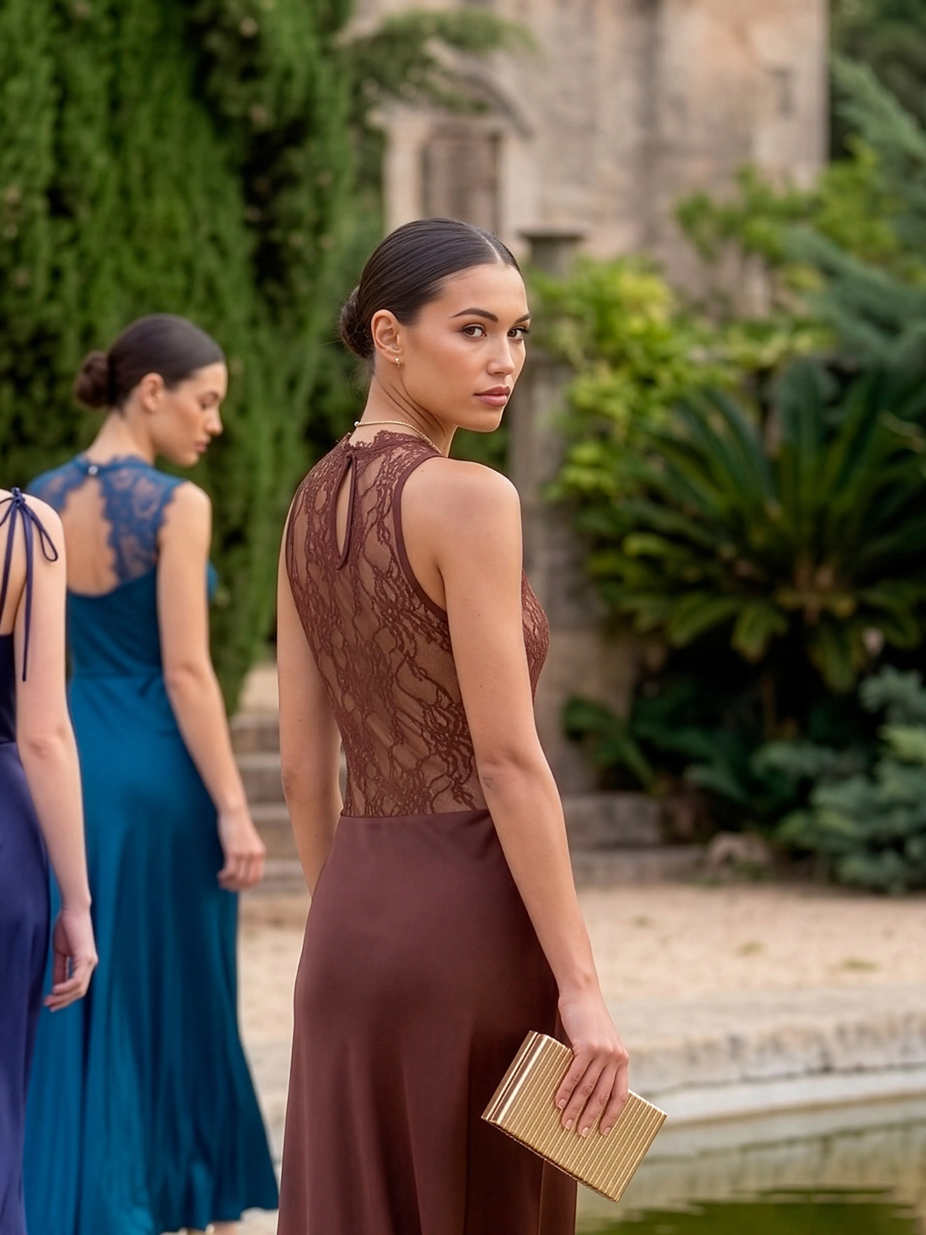 Three women in elegant evening gowns walk near a lush garden. The woman in the foreground turns her head back, holding a clutch, conveying an air of mystery.