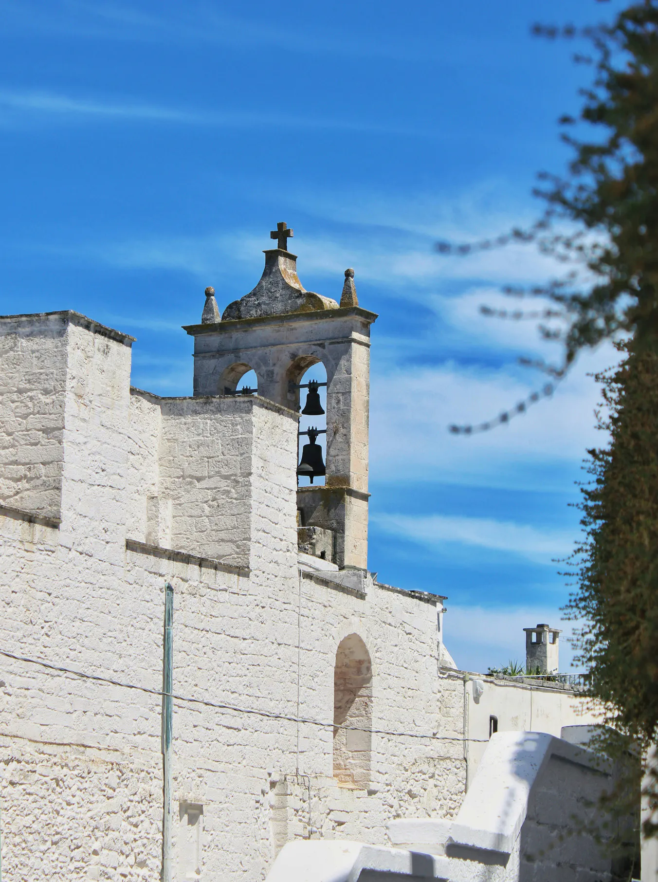 Stone church with two bells in an arched tower against a vibrant blue sky. The white walls and cross evoke a serene and timeless atmosphere.