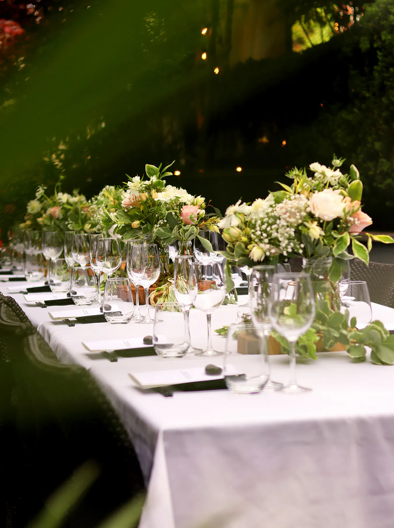 Elegant outdoor dining table with white cloth, adorned with lush floral centerpieces, crystal glassware, and place settings, creating a serene, festive atmosphere.