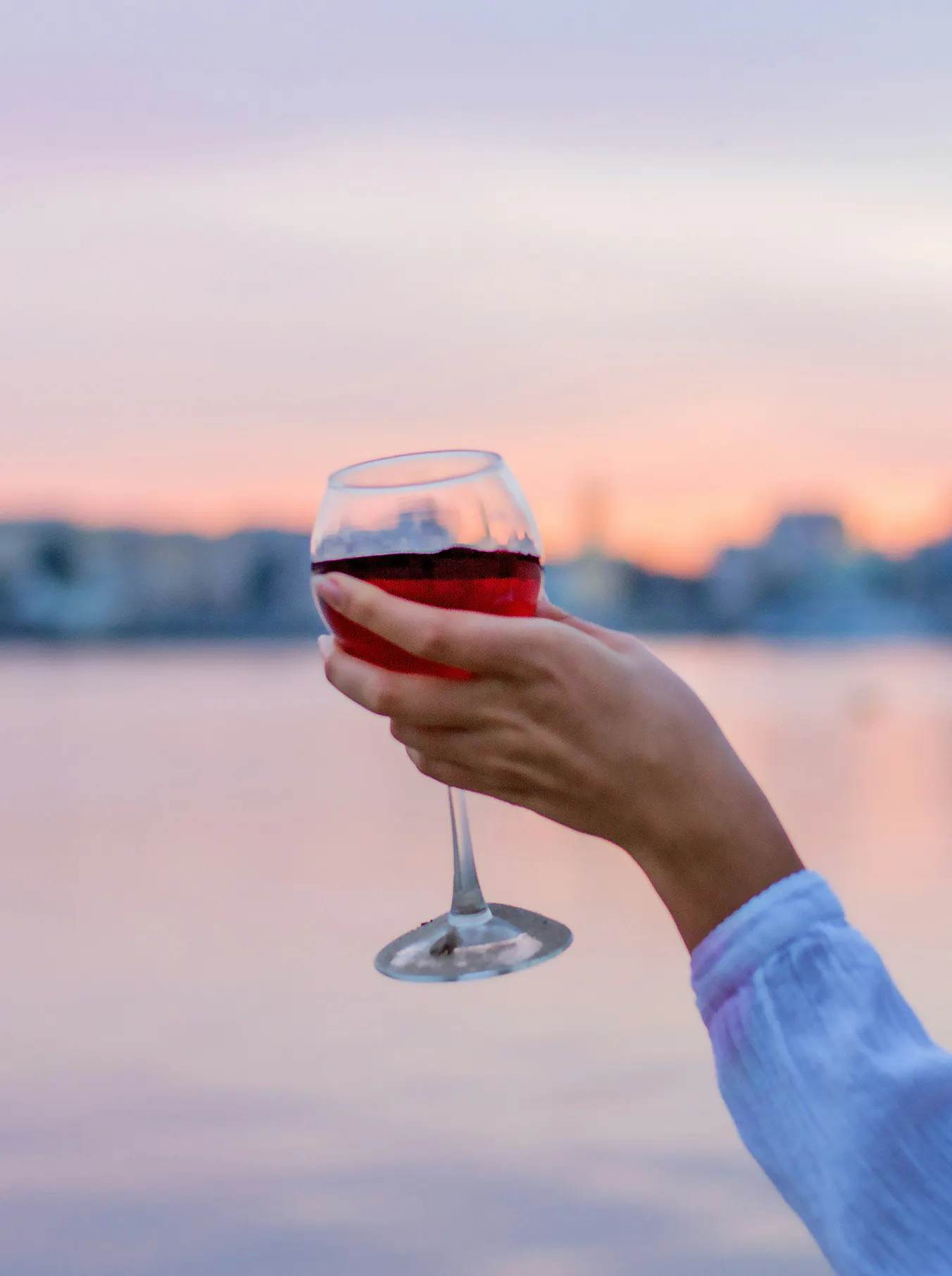 A hand holds a glass of red wine against a serene sunset. The sky is painted in soft pink and orange hues, reflecting a calm and relaxing atmosphere.